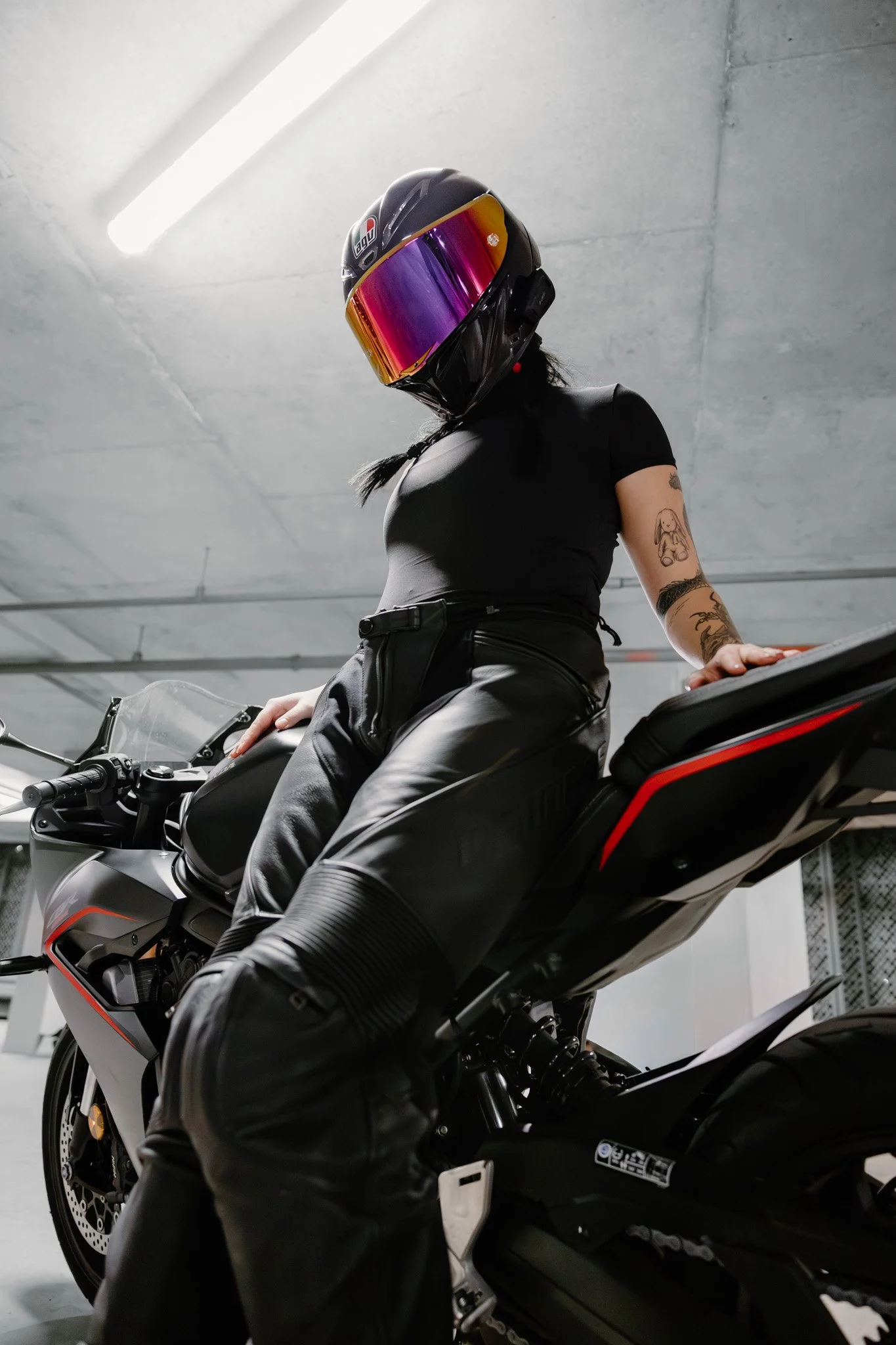 Person wearing a black helmet with a reflective visor, black t-shirt, and black leather pants sitting on a sport motorcycle in an indoor parking garage.