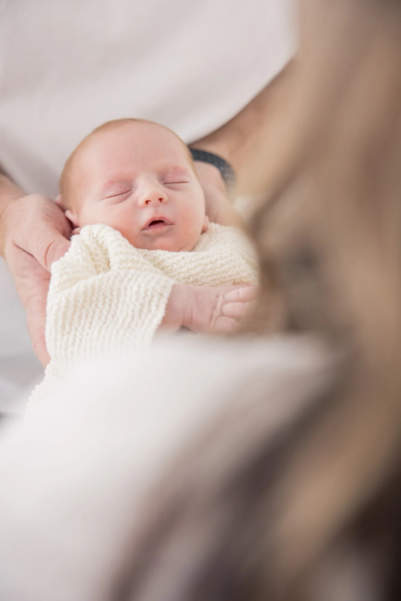 A newborn baby peacefully sleeping in a person's hands, wrapped in a cream-colored knitted blanket.