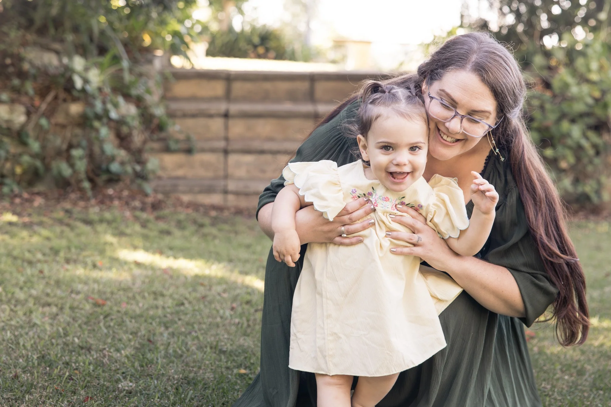 An older woman with glasses and long brown hair hugging a young girl in a yellow dress outdoors, with trees and sunlight in the background.