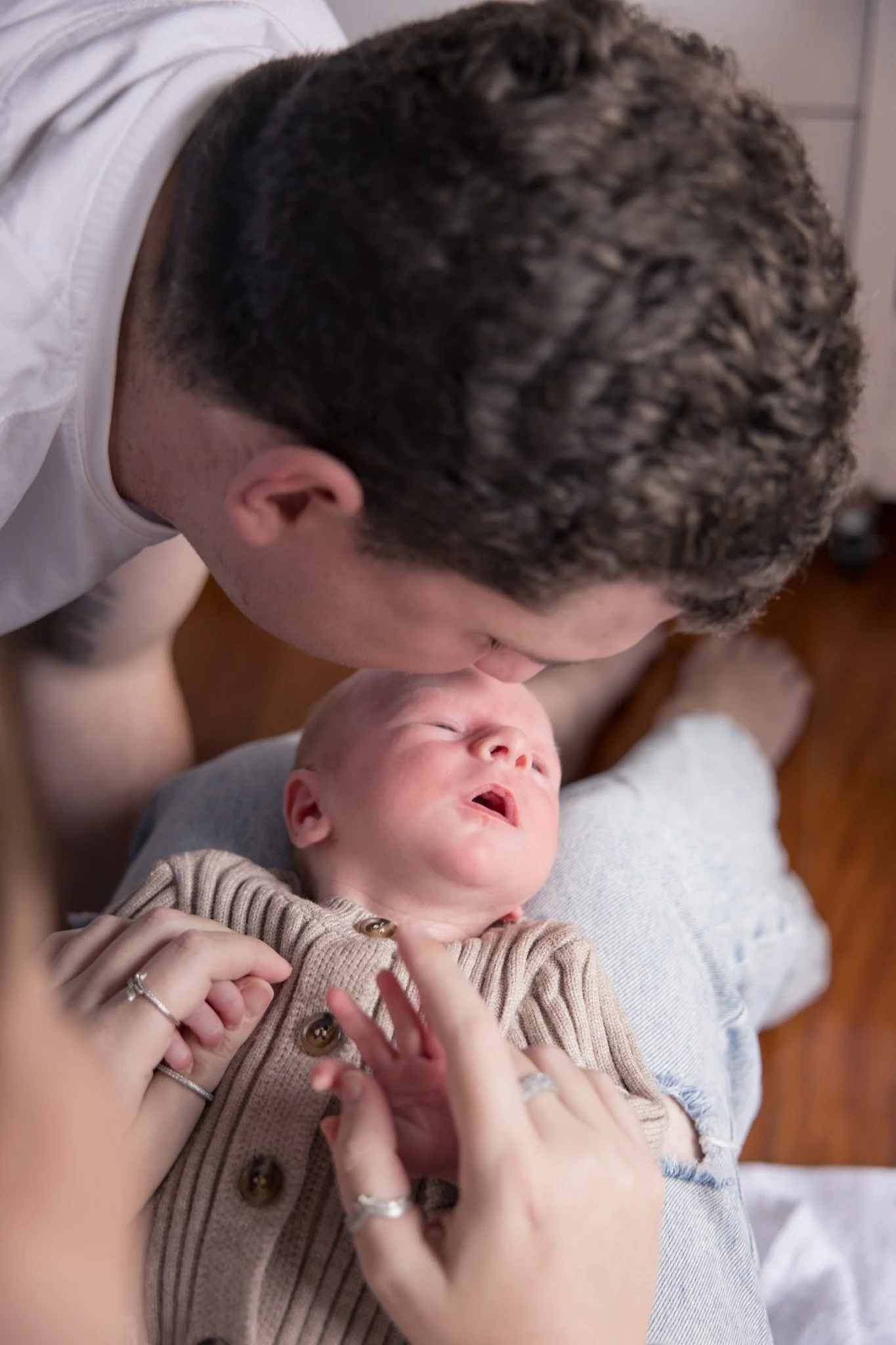 A man leaning over a baby lying on a person's lap, touching the baby's hands and face, in a warm indoor setting.