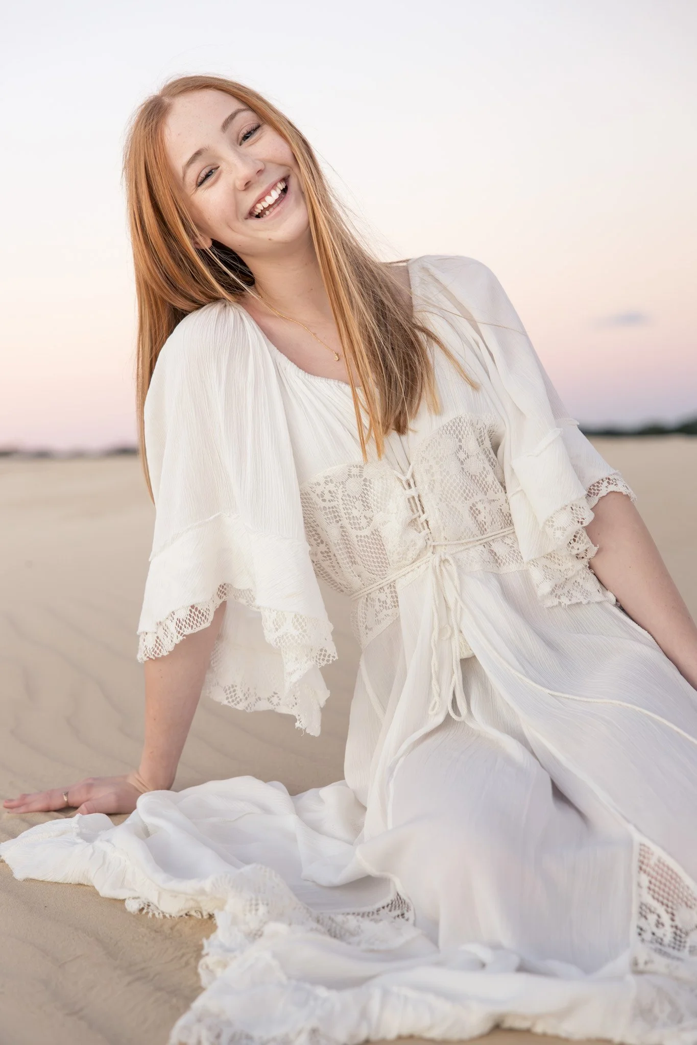 A young woman with red hair, smiling and sitting on a sandy beach during sunset or sunrise, wearing a white dress with lace details.
