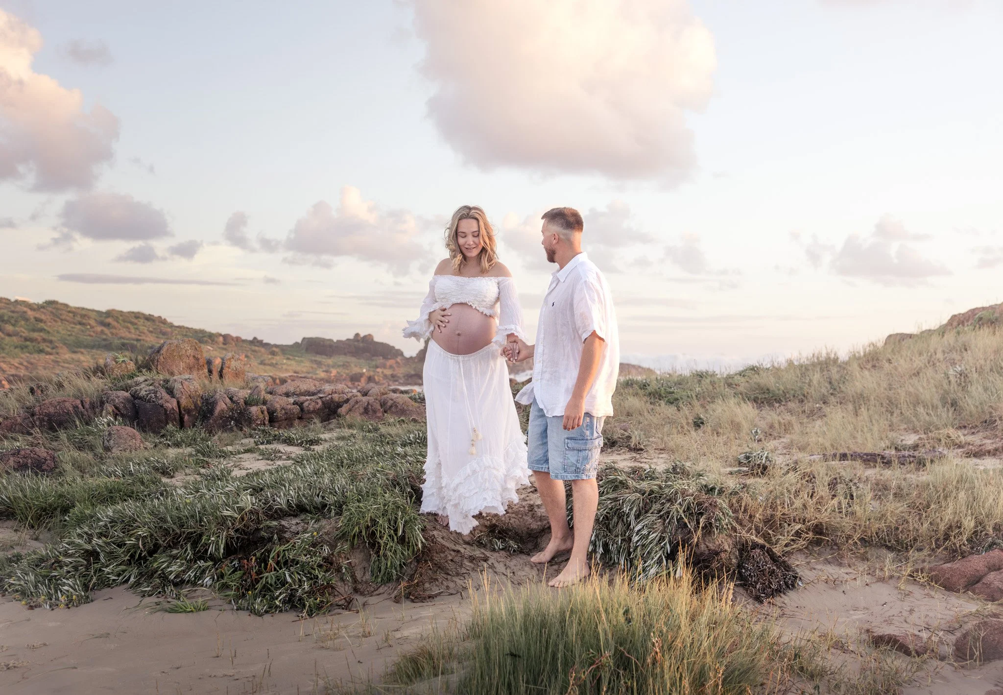A pregnant woman in a white dress holding her belly and a man holding her hand, standing barefoot on a beach with rocks and grass, under a cloudy sky at sunset. Photo taken at anna bay beach nsw