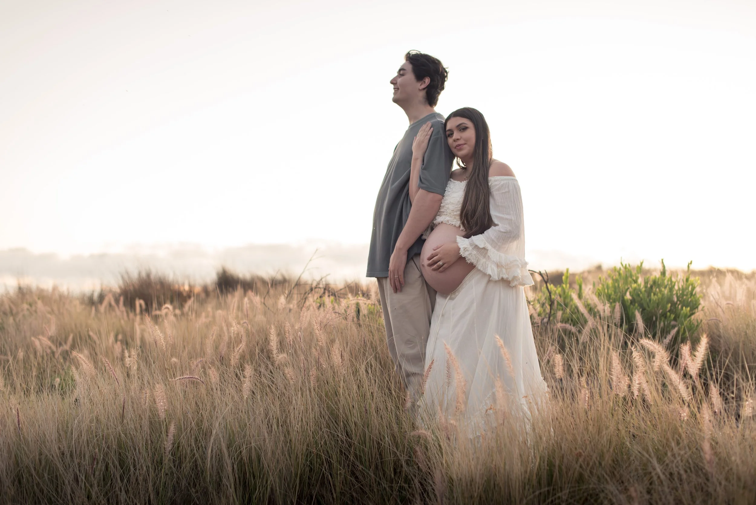 A pregnant woman in a white dress and a young man in a gray shirt stand in a grassy field during sunset, with the woman resting her hand on her belly and the man smiling with his eyes closed.
