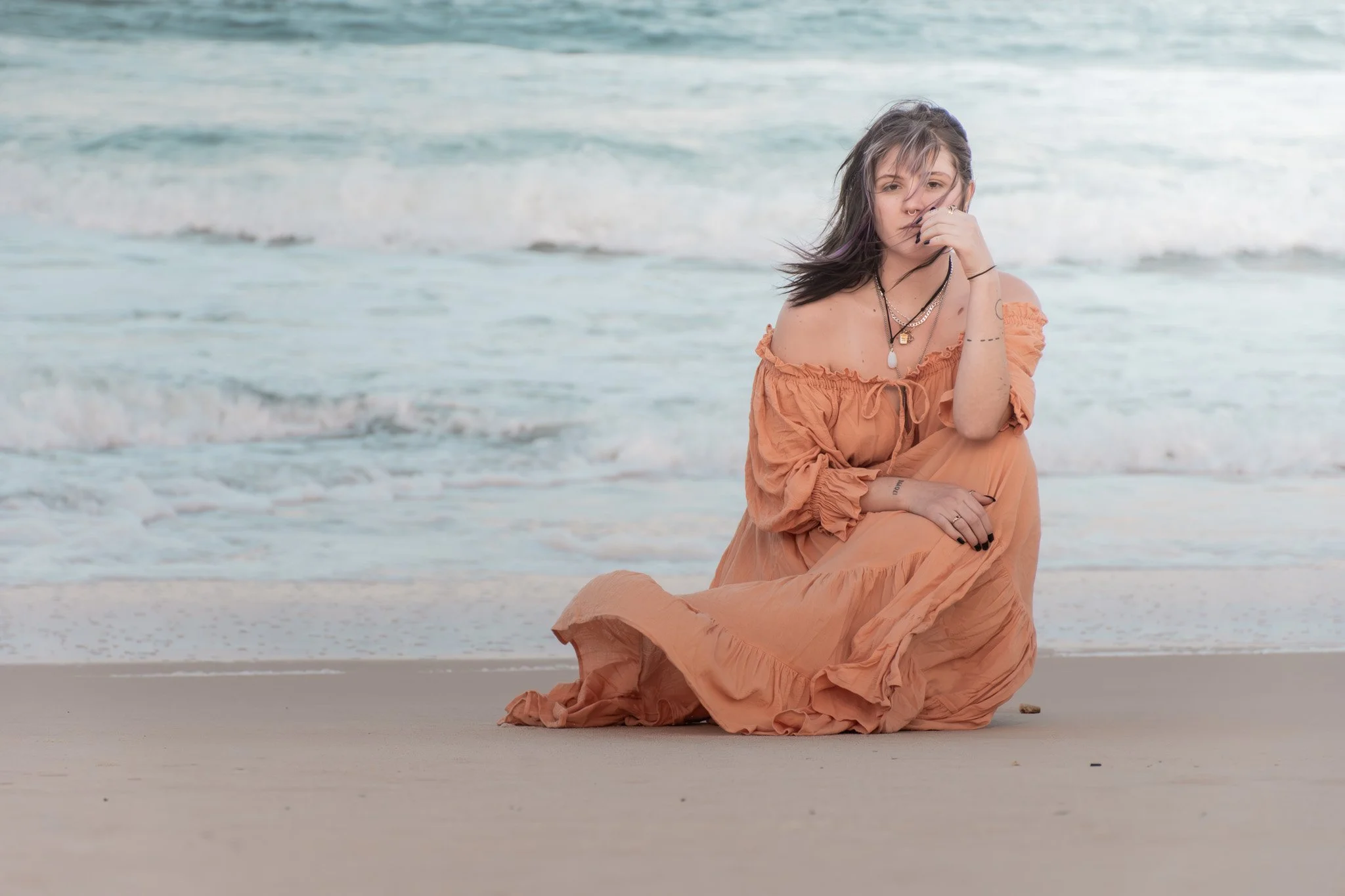 A woman sitting on the beach near the water, wearing an orange off-shoulder dress, with long dark hair, and layered necklaces.