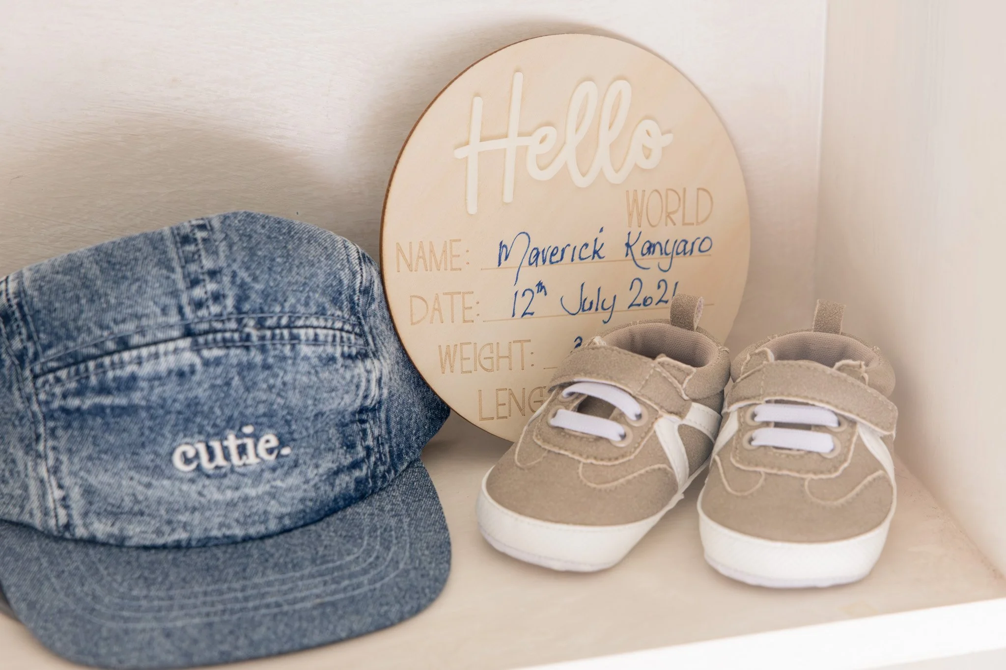 A wooden plaque with the words 'Hello World' and handwritten details about a baby, including name Maverick Kangaro, date of birth July 12, 2021, and baby shoes beside a denim hat with the word 'cutie' embroidered on it.