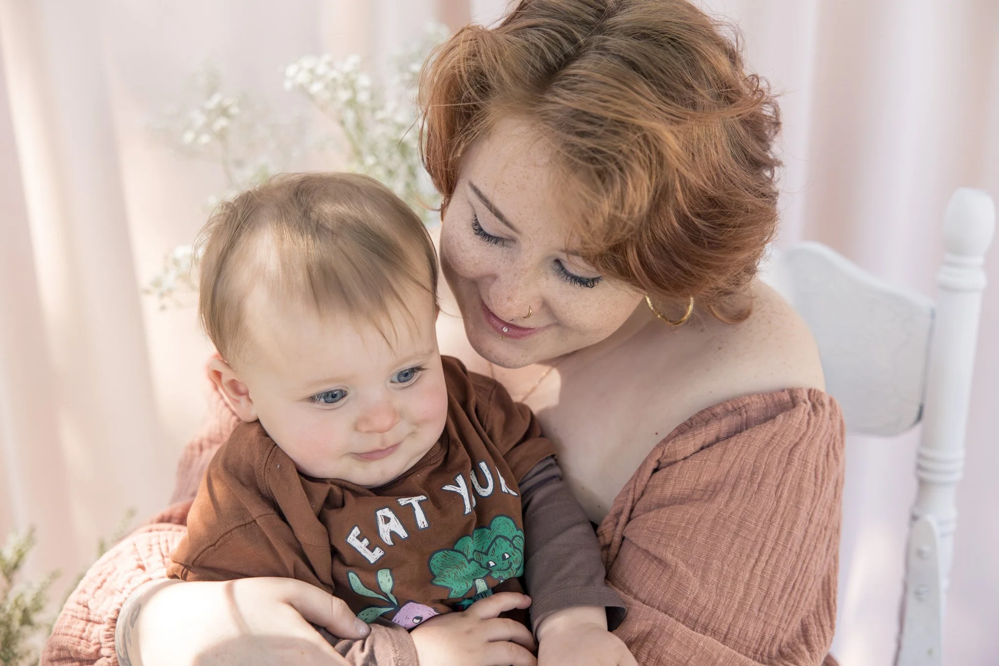 A woman with red hair, freckles, gold hoop earrings, and a nose ring is hugging a young boy with blonde hair and blue eyes. The woman is smiling and the boy has a neutral expression. They are sitting indoors near a white bed with a white headboard, with soft natural light and a background of light-colored curtains and floral decorations.