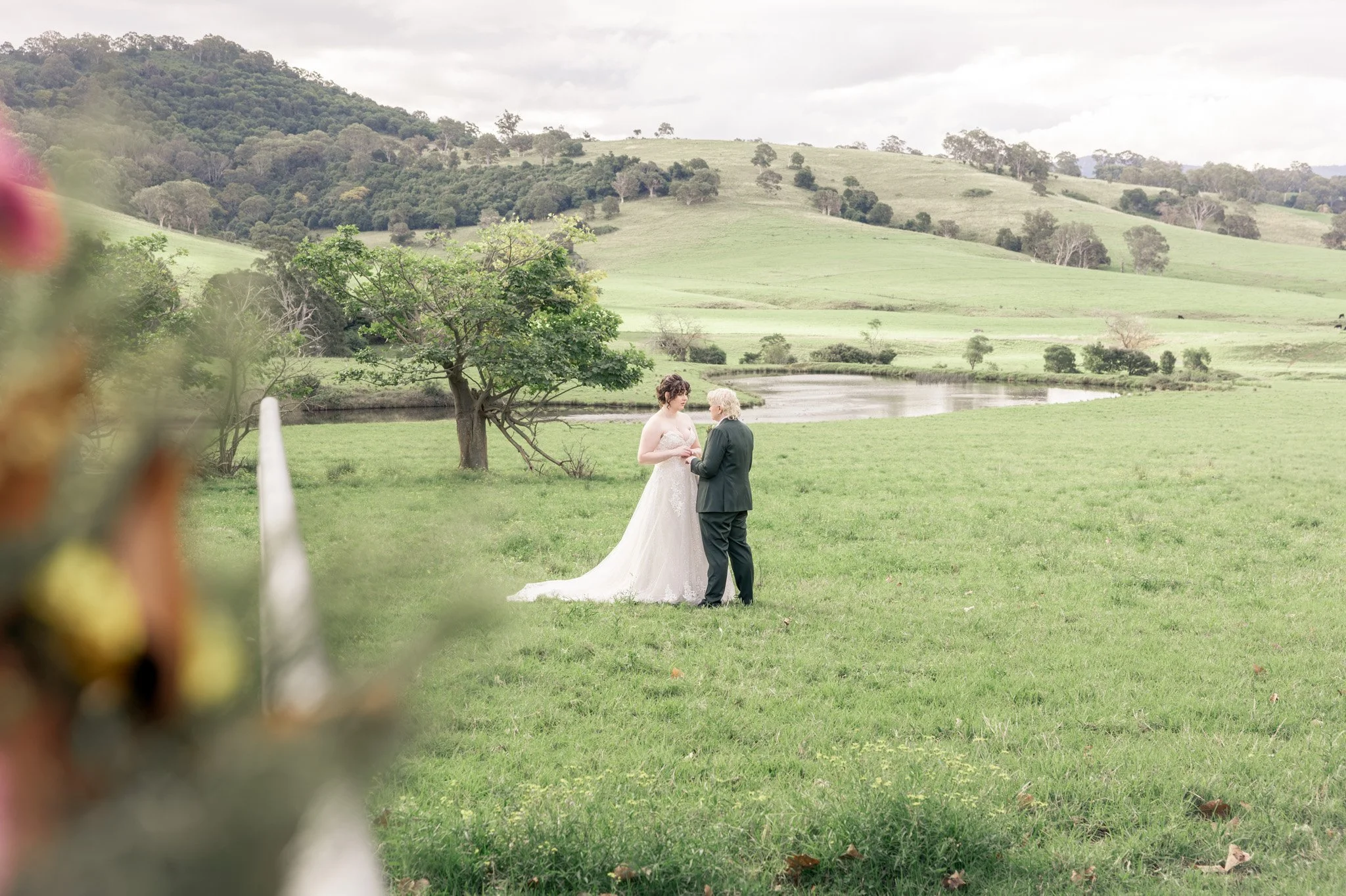 A bride and groom standing together outdoors on a grassy area near a pond, with green hills and trees in the background, during a wedding ceremony.