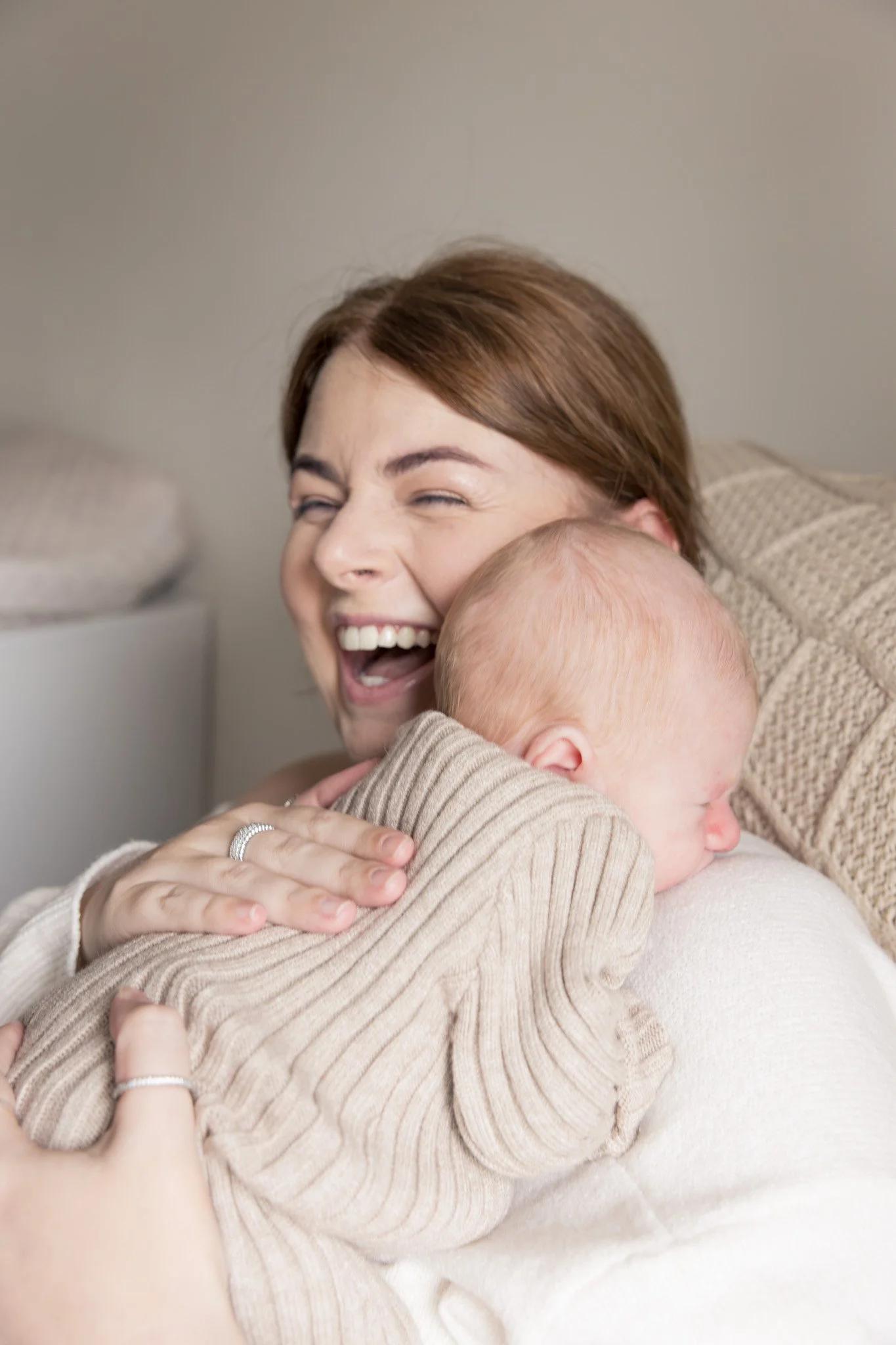 A woman with brown hair smiling and laughing while hugging a baby with blonde hair, who is facing away from the camera. They are dressed in light-colored, cozy clothing and appear to be enjoying a joyful moment together indoors.