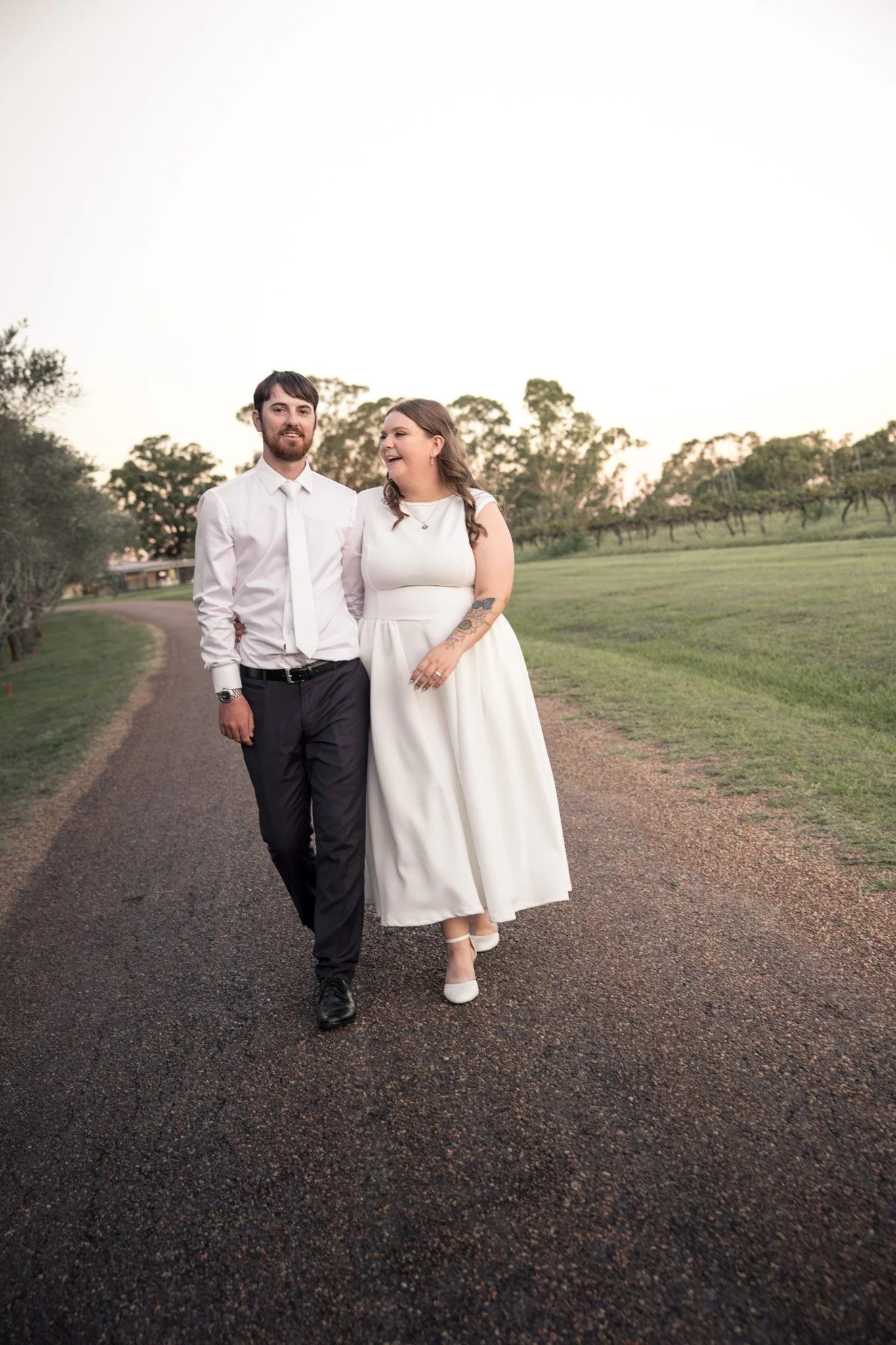 A couple dressed in wedding attire walking hand-in-hand outdoors on a paved path, smiling and looking at each other, with trees and a light sky in the background.
