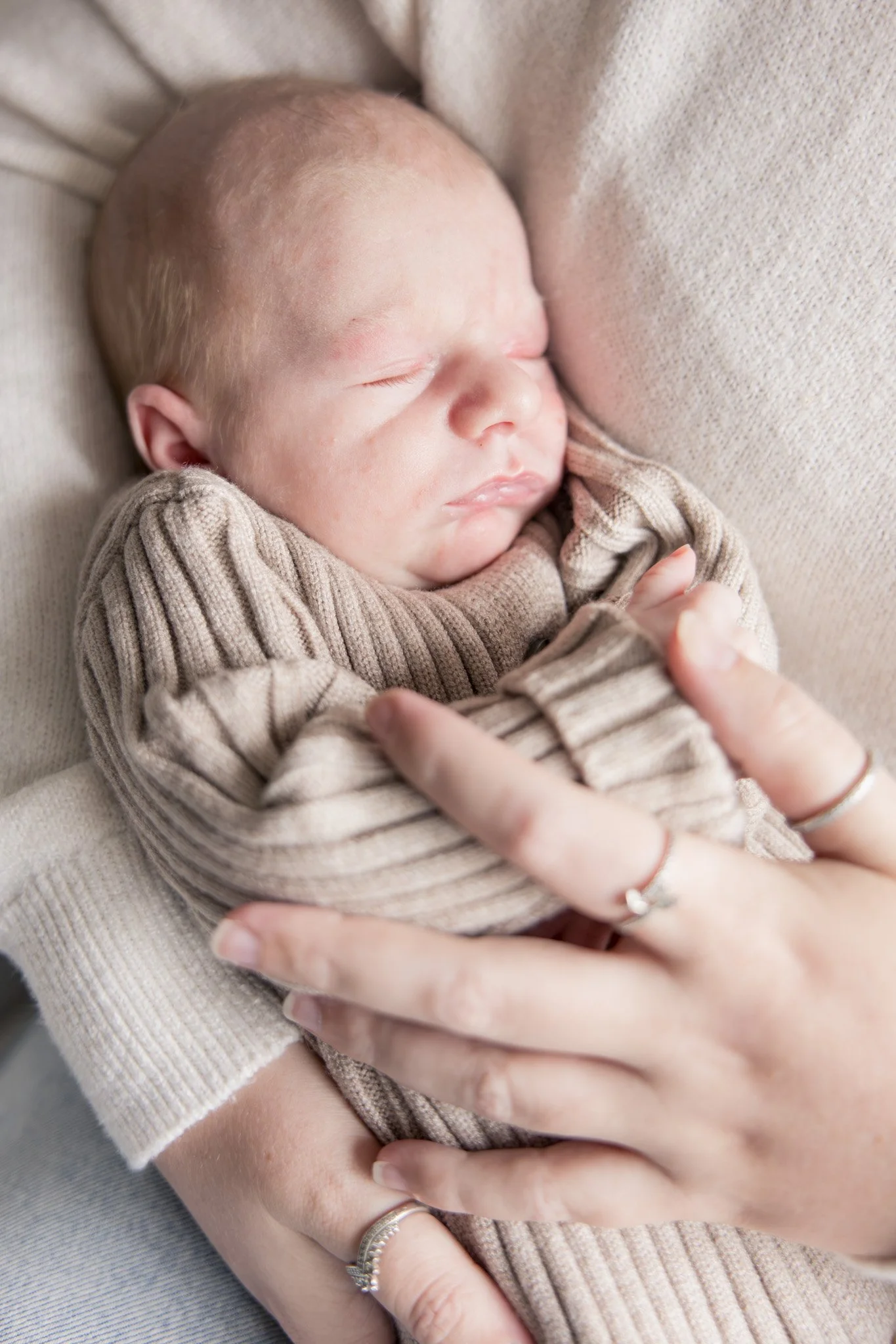 A sleeping baby with light-colored hair wearing a beige striped sweater rests. An adult's hands gently cradle the baby's body, an adult's hand with rings is visible in the image.
