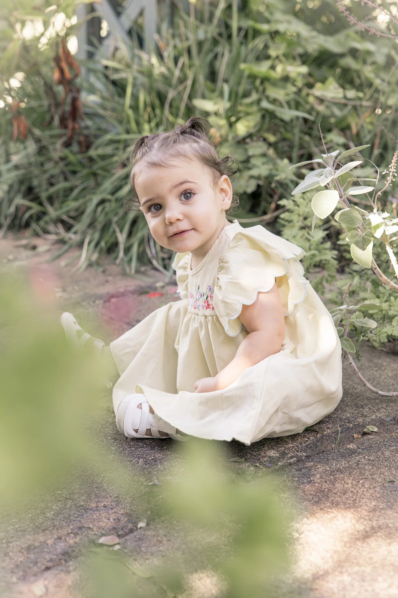 A young girl with curly hair and big blue eyes sitting on the ground outdoors, surrounded by green plants and foliage, wearing a light yellow dress with colorful embroidery.