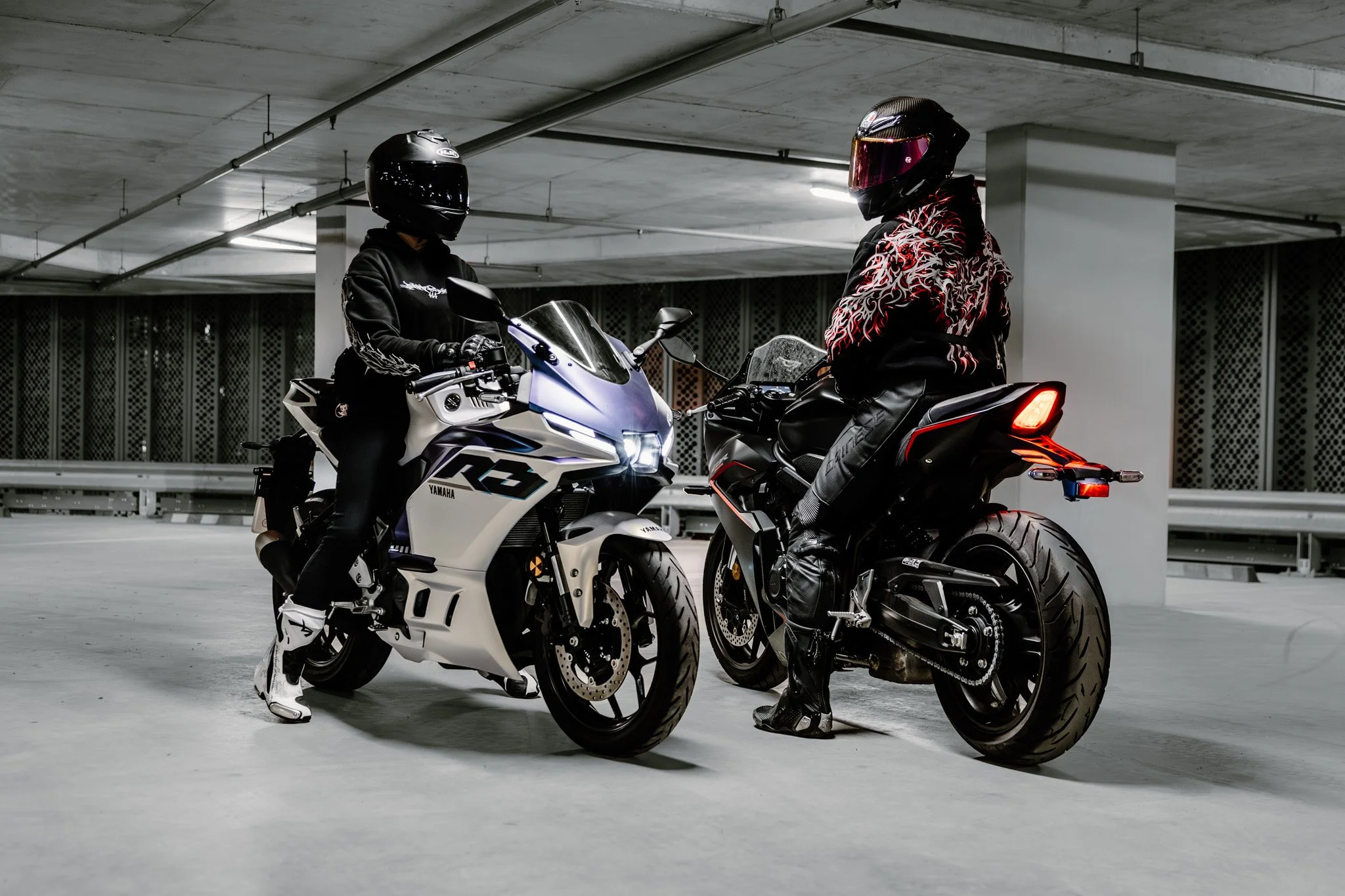Two motorcyclists in full gear, including helmets, standing next to their motorcycles in an indoor parking garage with concrete ceilings and floors.