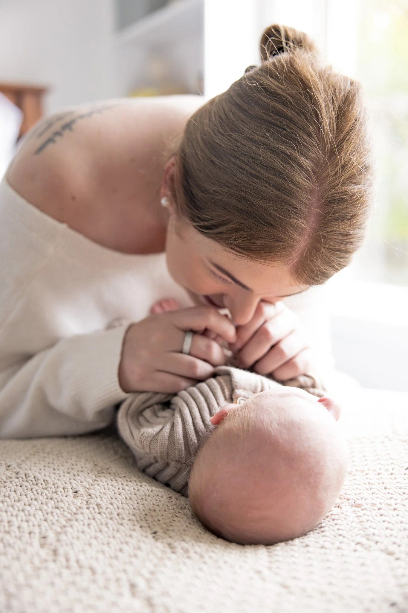 A woman with light brown hair leaning over and kissing a baby who is lying on a beige carpet, with soft lighting in the background.