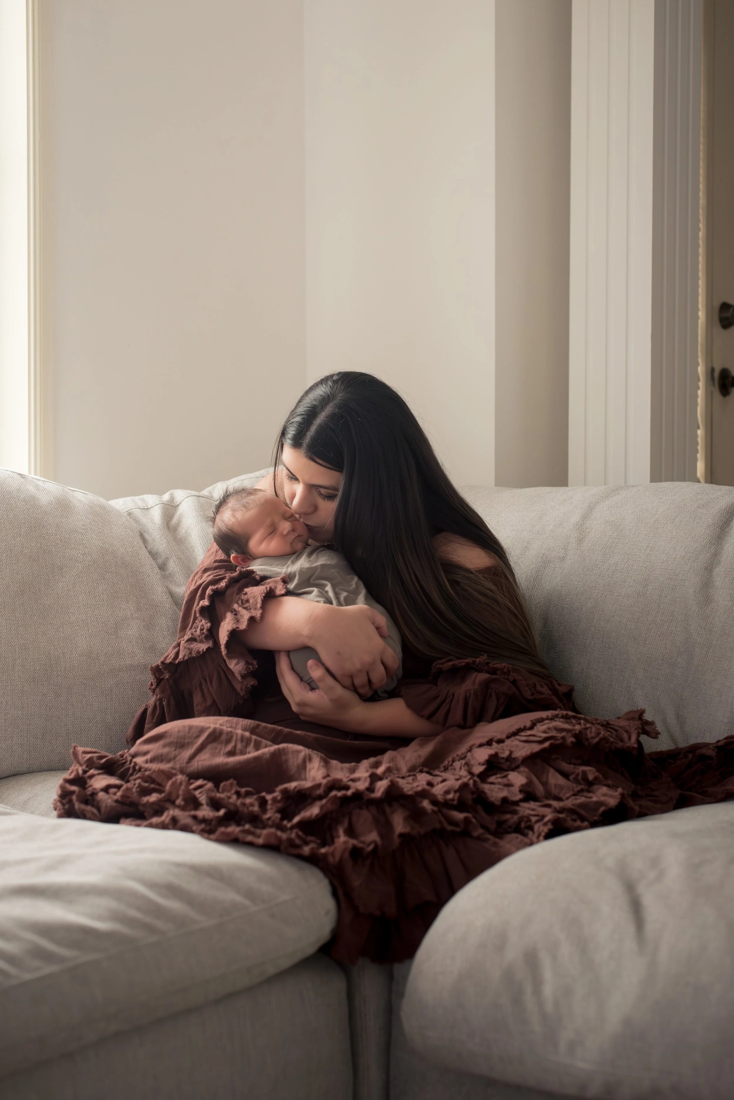 A woman with long dark hair sitting on a beige couch, holding a sleeping baby wrapped in a light-colored blanket, in a room with light-colored walls.