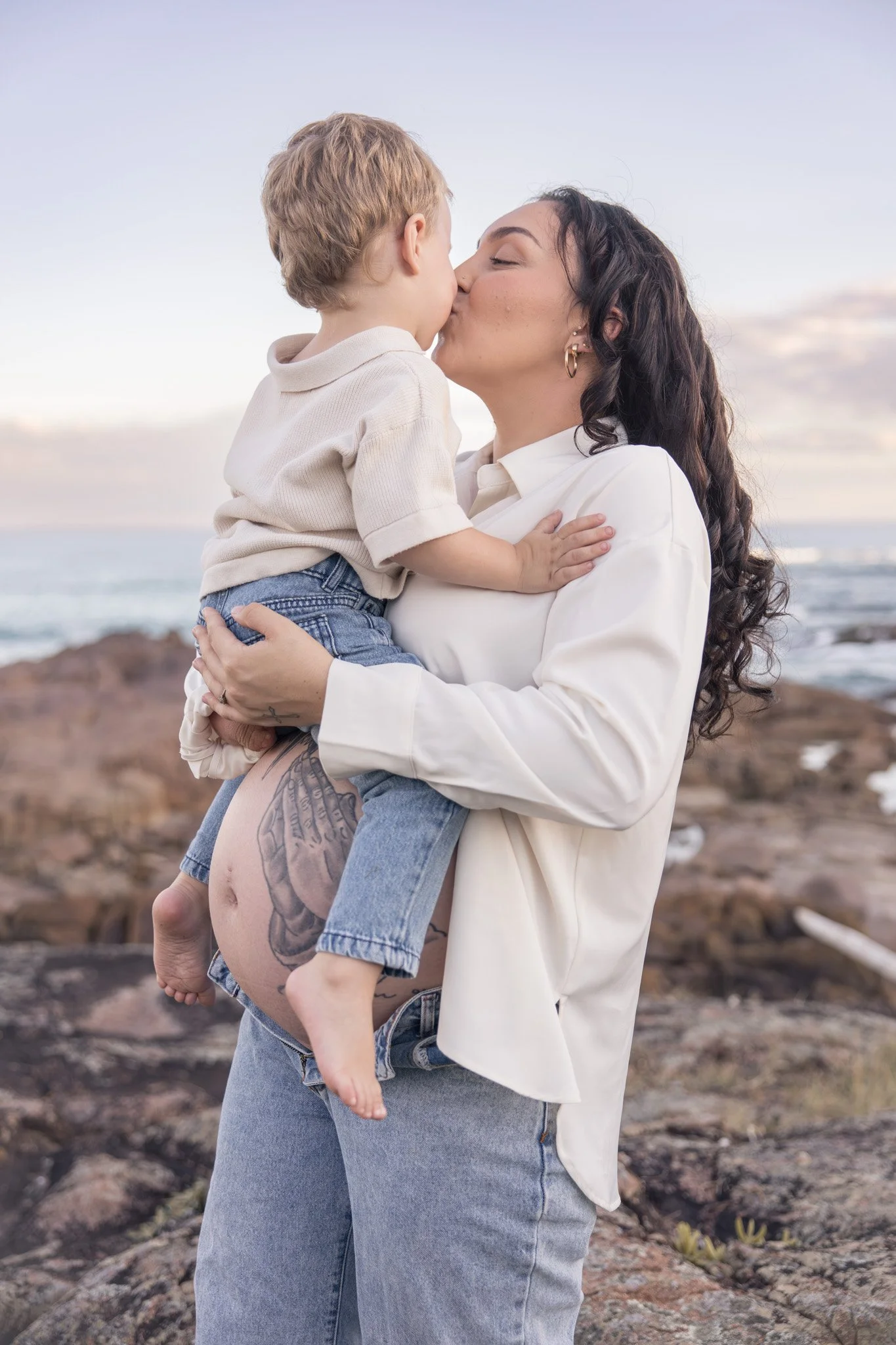 A woman with dark hair and tattoos on her leg holding a young boy with blonde hair as they kiss with the ocean and rocks in the background.