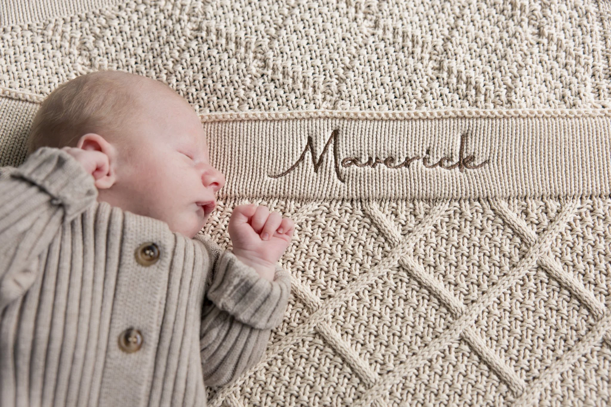 A sleeping baby with light hair wearing a beige striped sweater, lying on a textured beige blanket with the word 'Mamá' embroidered on it.