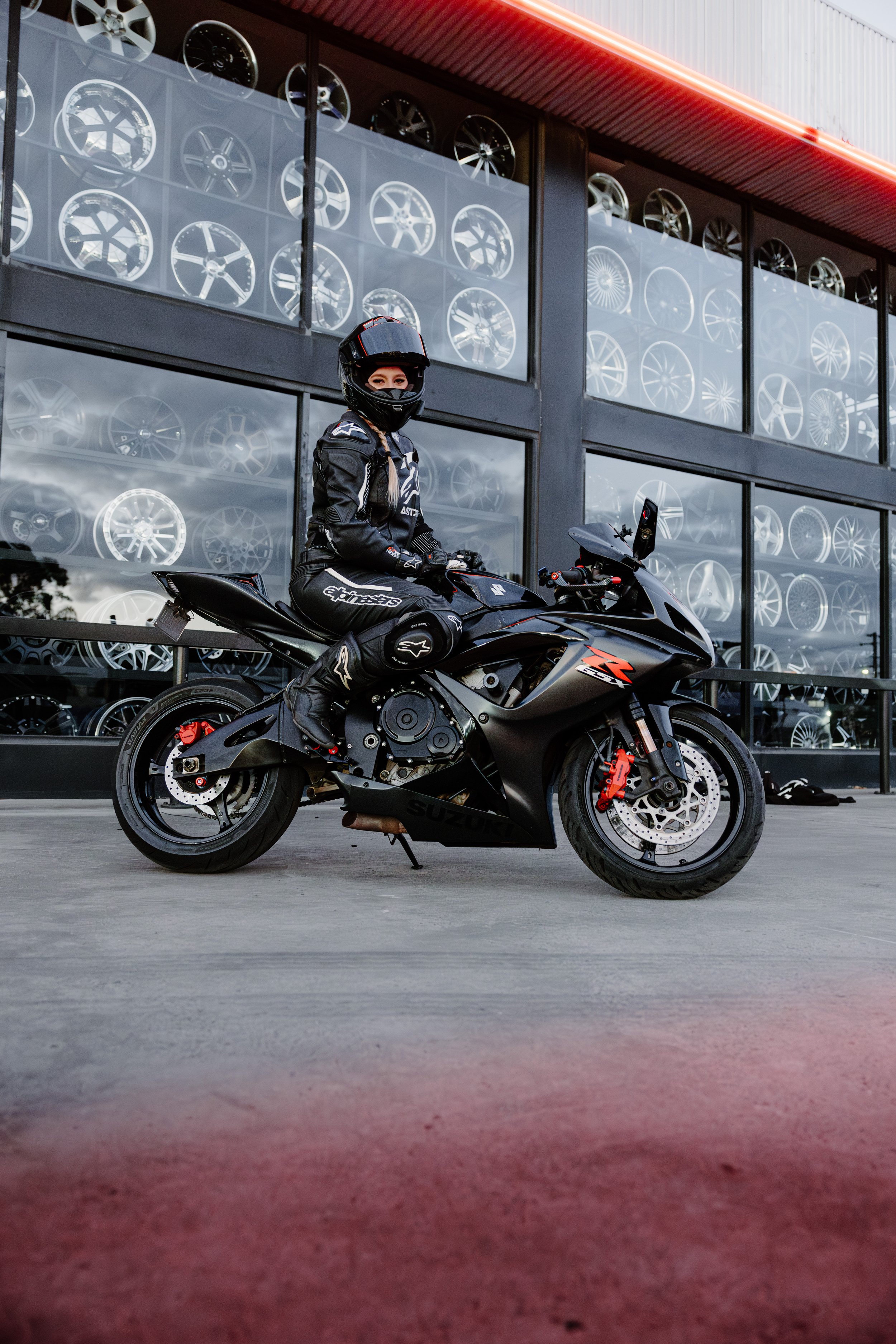 A woman wearing motorcycle gear, including a helmet, sitting on a black Suzuki sport motorcycle outside a storefront displaying alloy wheels in the windows.