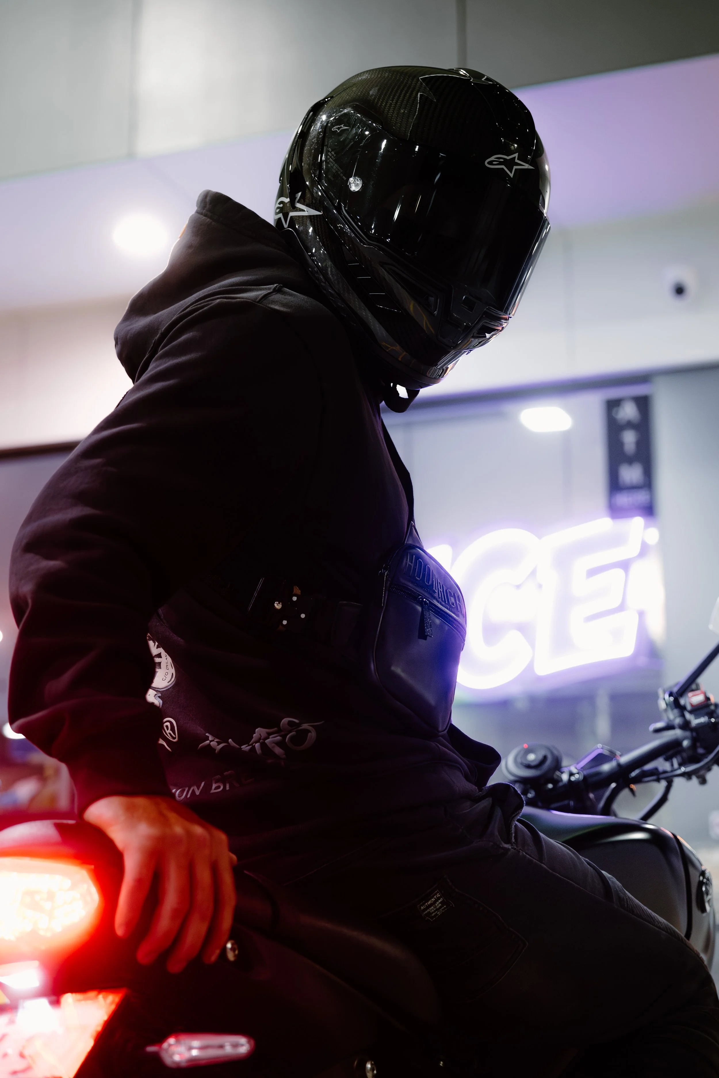 A person in a black motorcycle helmet and dark clothing sitting on a red motorcycle, with their left hand visible near the tail light. The background has a neon sign and indoor lighting.