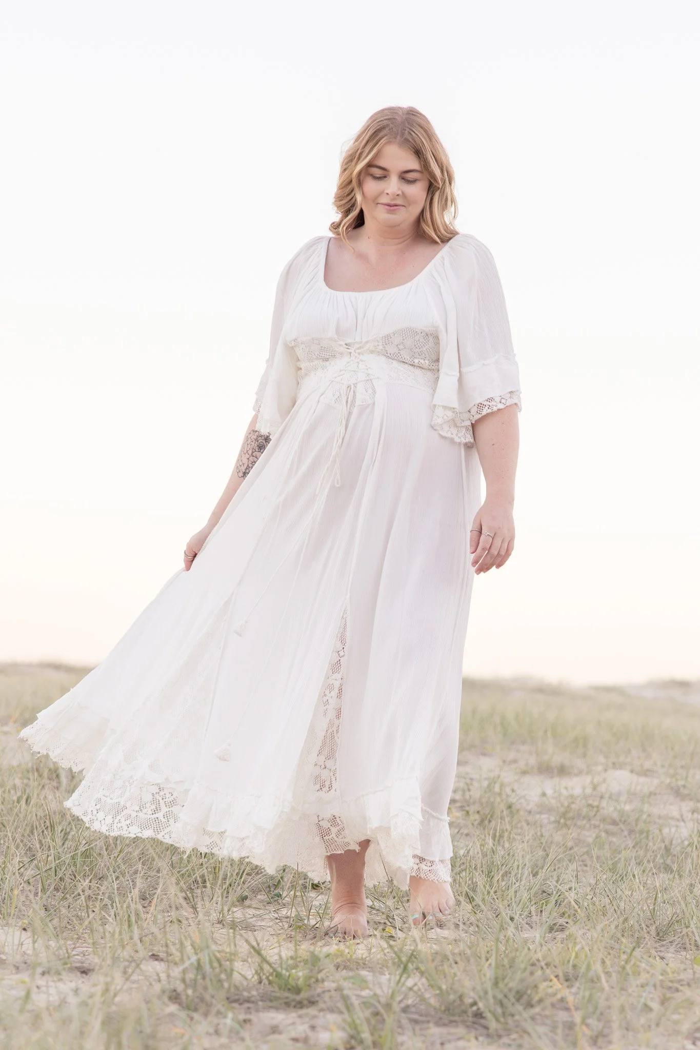 A woman in a flowing white dress walking barefoot on a grassy field, with a light and airy background. Photo taken at lighthouse beach port maquiarie