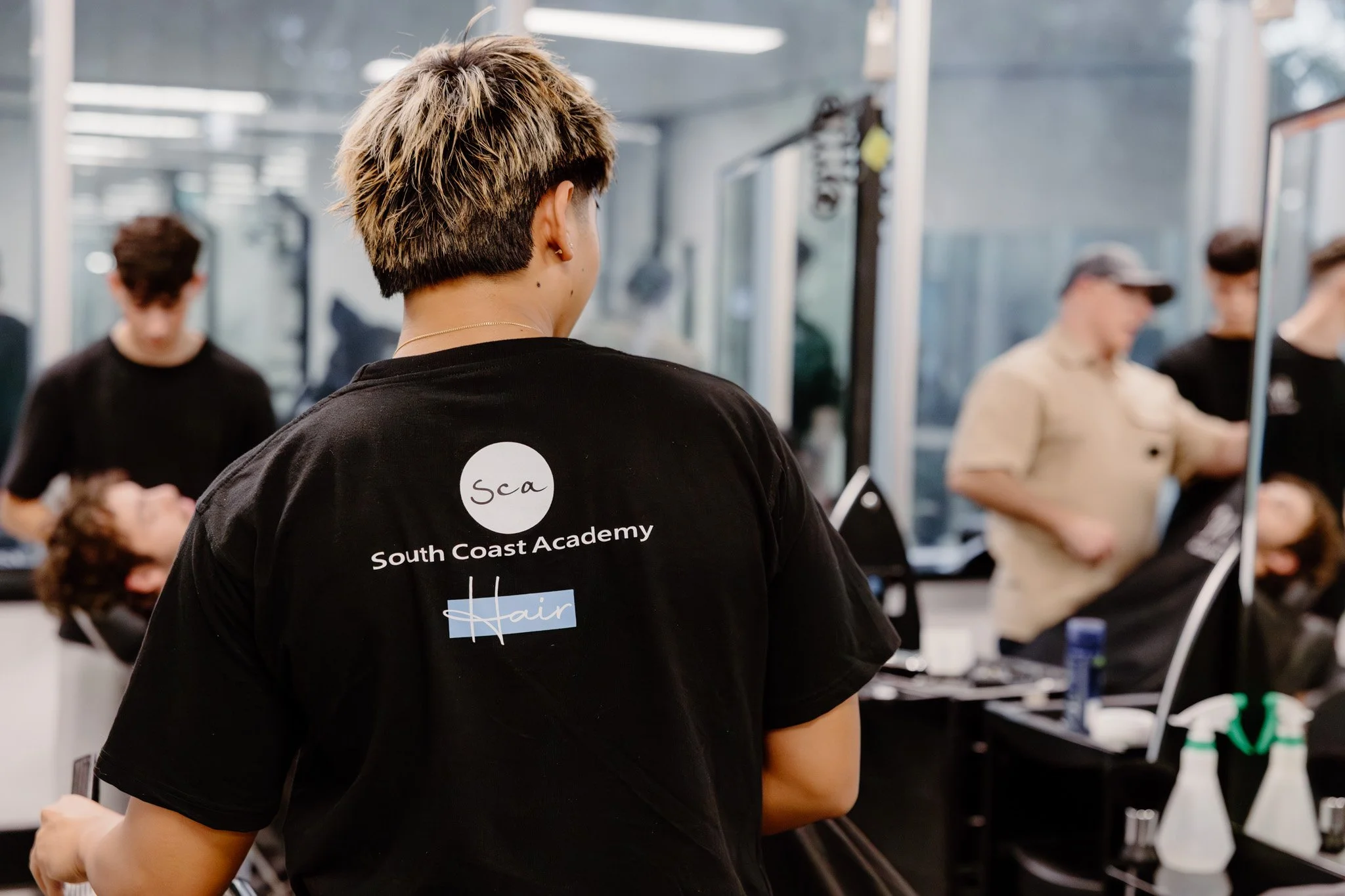 A person with short, layered hair with blonde highlights, wearing a black t-shirt with a sticker that says "SCA South Coast Academy Hair" on the back, stands in a salon or barbershop surrounded by other people, some getting haircuts or styling.
