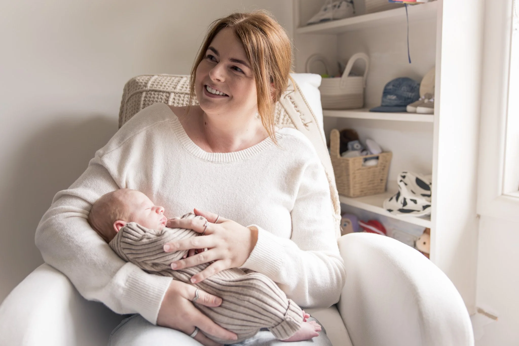 A woman with red hair and a white sweater sitting on a white armchair, smiling while holding a baby wrapped in a striped blanket. In the background, there are shelves with folded clothes, a basket, and a window with light coming through.