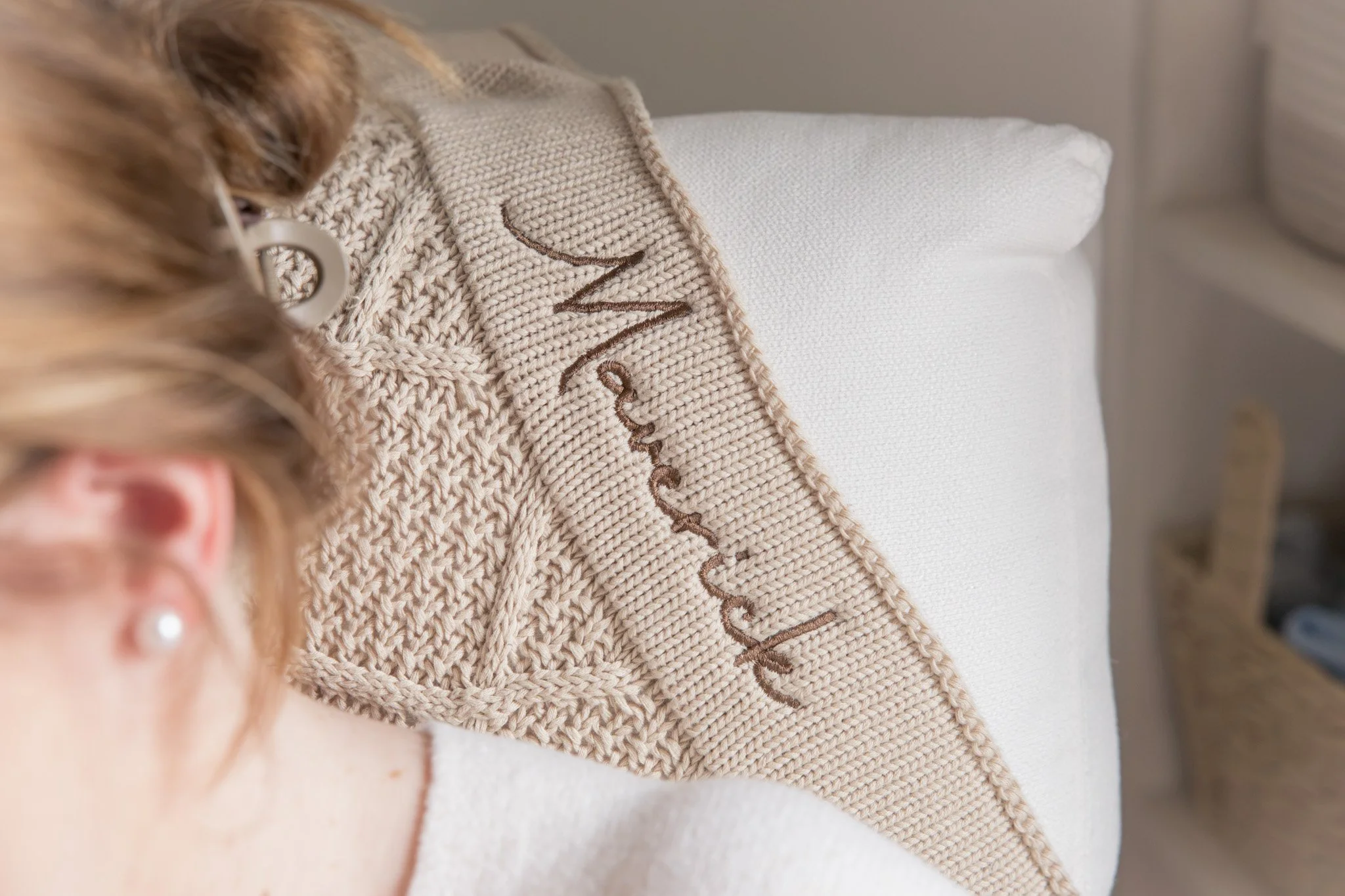 Close-up of a knitted beige blanket with embroidered brown script, draped over a white textured surface, with a blurred glimpse of a person's hand and jewelry in the foreground.