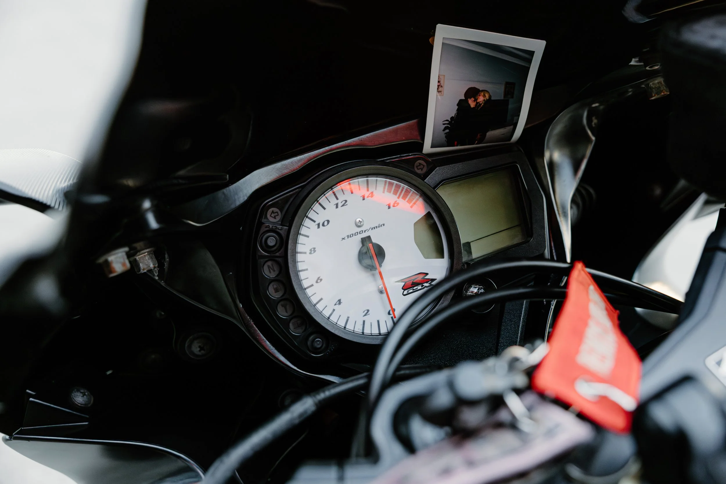 Close-up of a motorcycle dashboard with a large white tachometer, a small digital display, and a red cloth or strap hanging nearby.