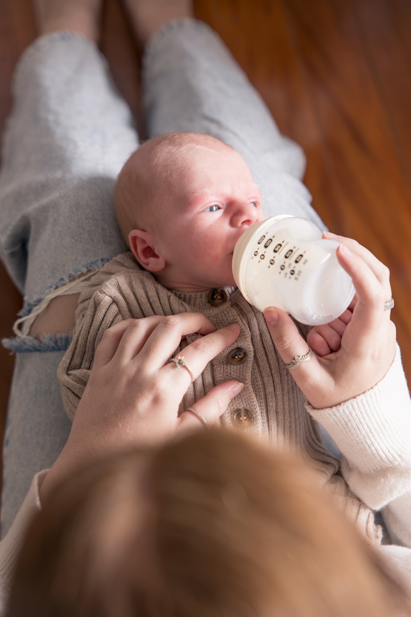 A baby with light skin and blue eyes lying on a person's lap, drinking from a baby bottle, with a wooden floor background.