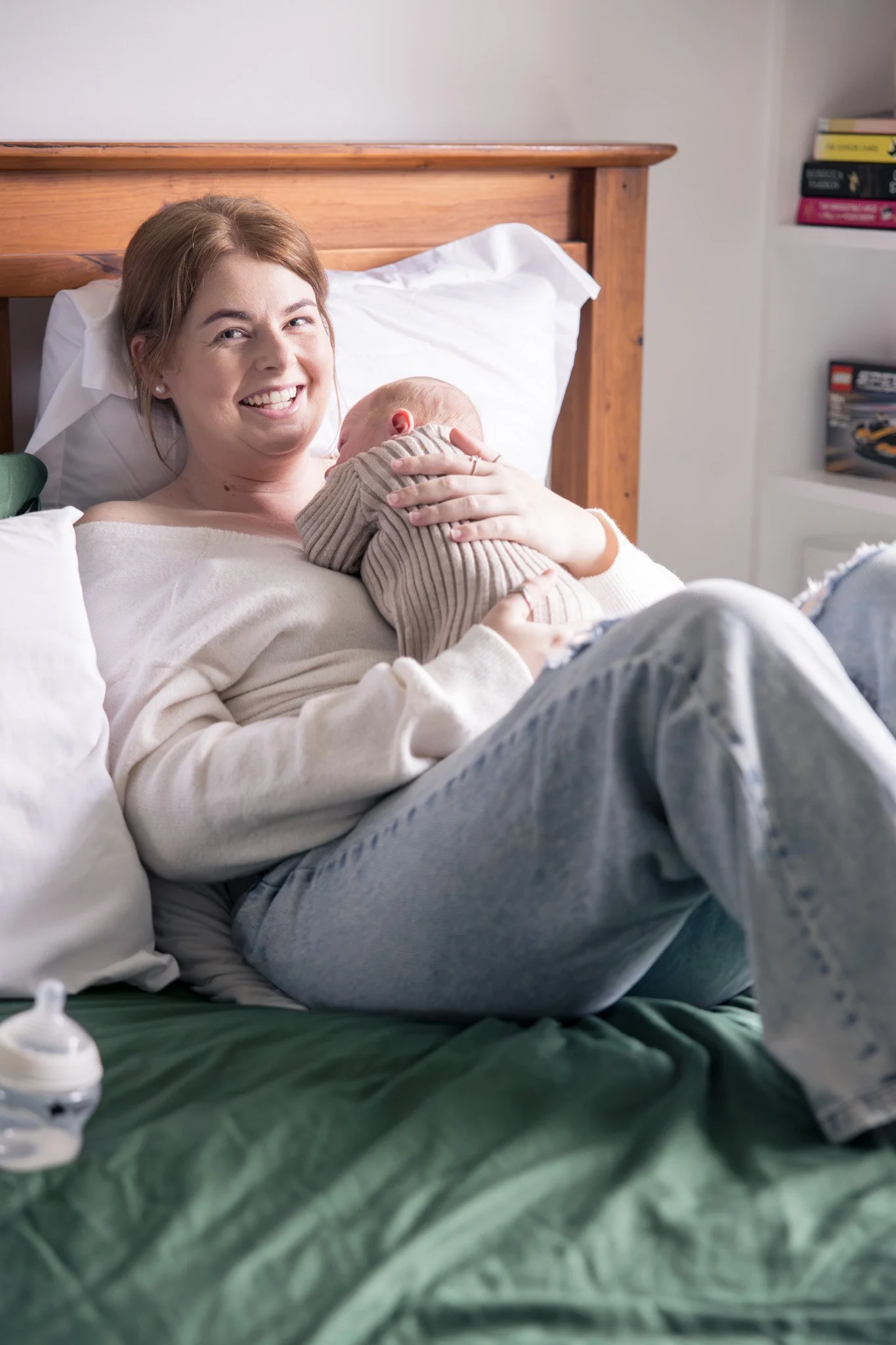 A woman with brown hair smiling while holding a newborn baby in a beige striped outfit in a bedroom.