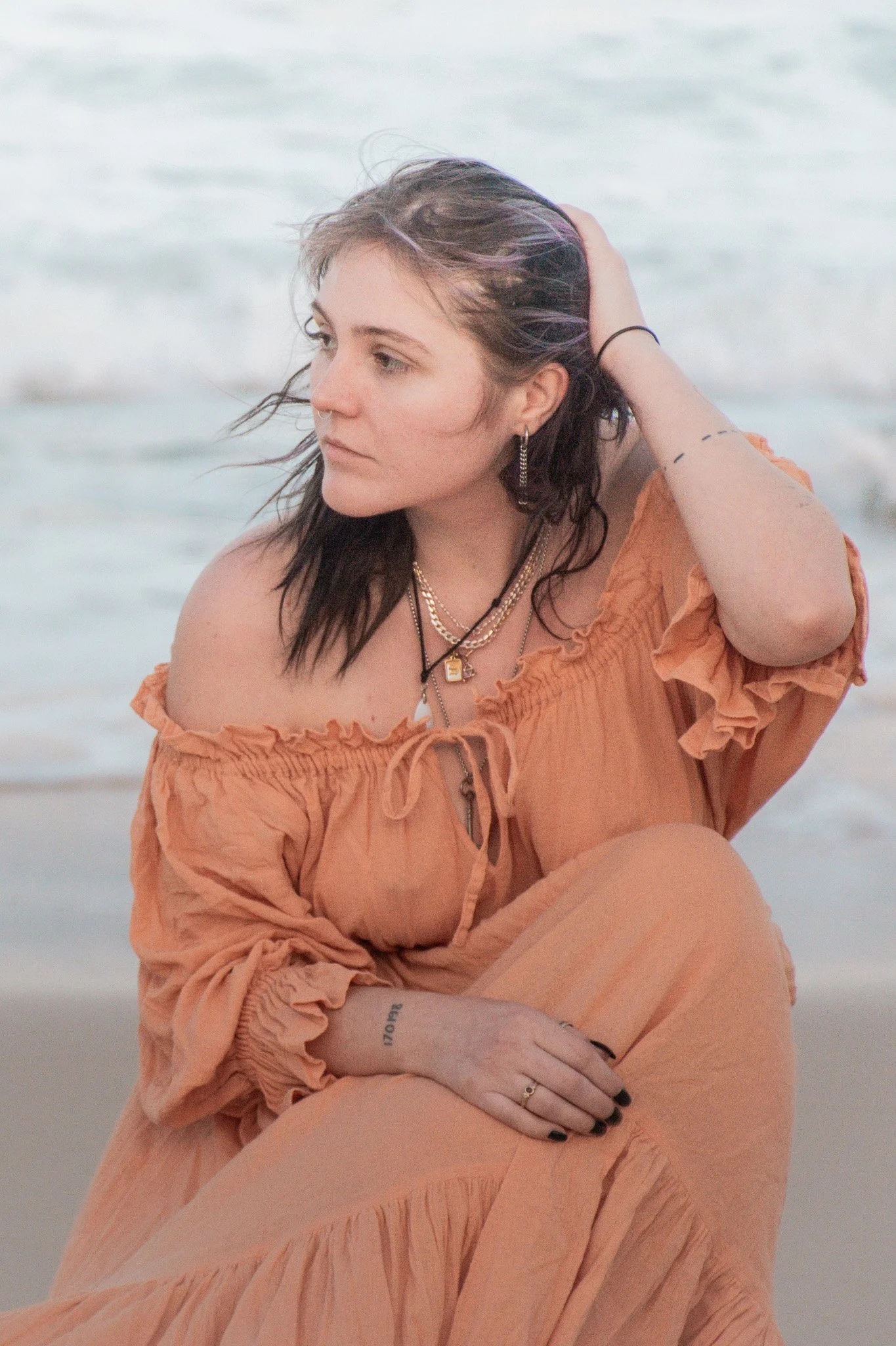 A woman with wavy dark hair poses on the beach, wearing an orange off-the-shoulder dress and layered necklaces, with her hand on her head and sitting near the shoreline.