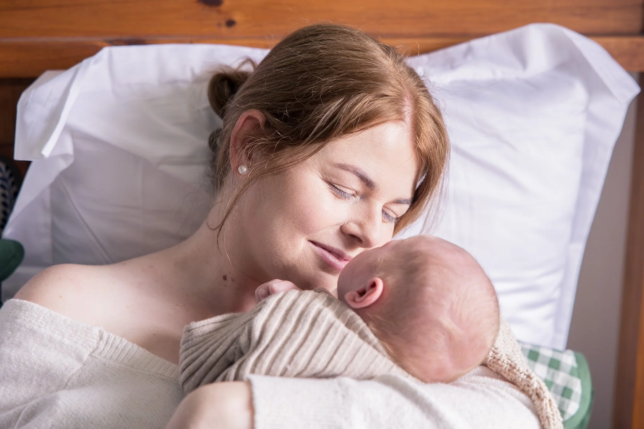 A woman with light brown hair lying in bed, smiling and kissing a baby's forehead, with a wooden headboard and white bedding visible.
