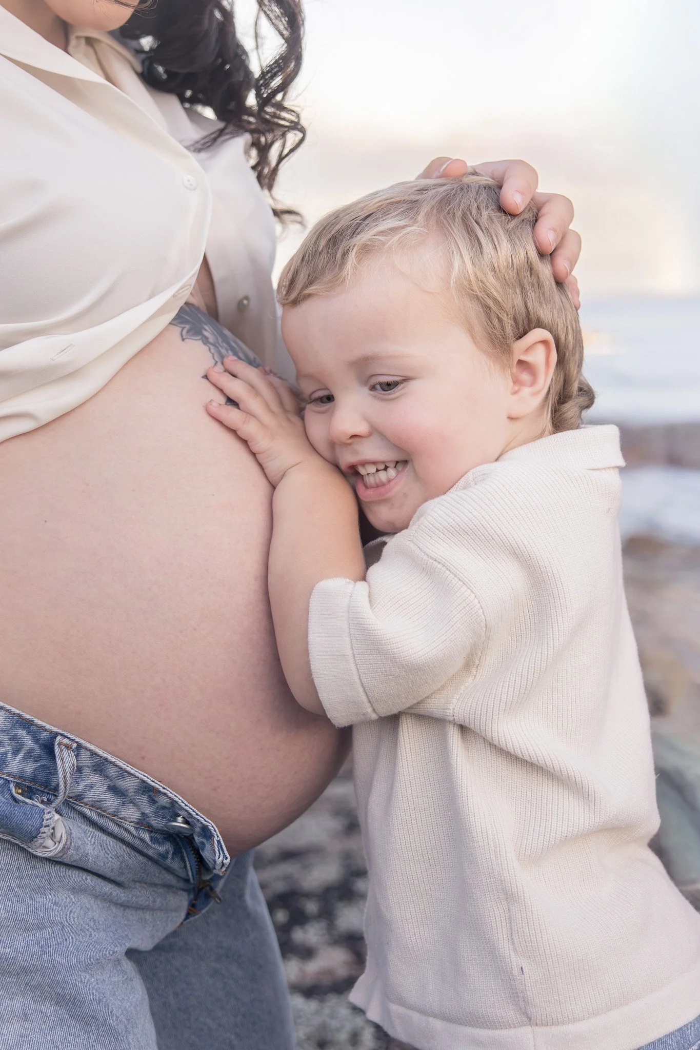A woman with long dark hair, wearing a cream-colored shirt, is standing close to a young boy with light-colored hair, who is smiling and hugging her pregnant belly. The boy's hand is resting on the pregnant belly, and the woman is gently holding the boy's head. They are outdoors at the beach, with sand and the ocean visible in the background.