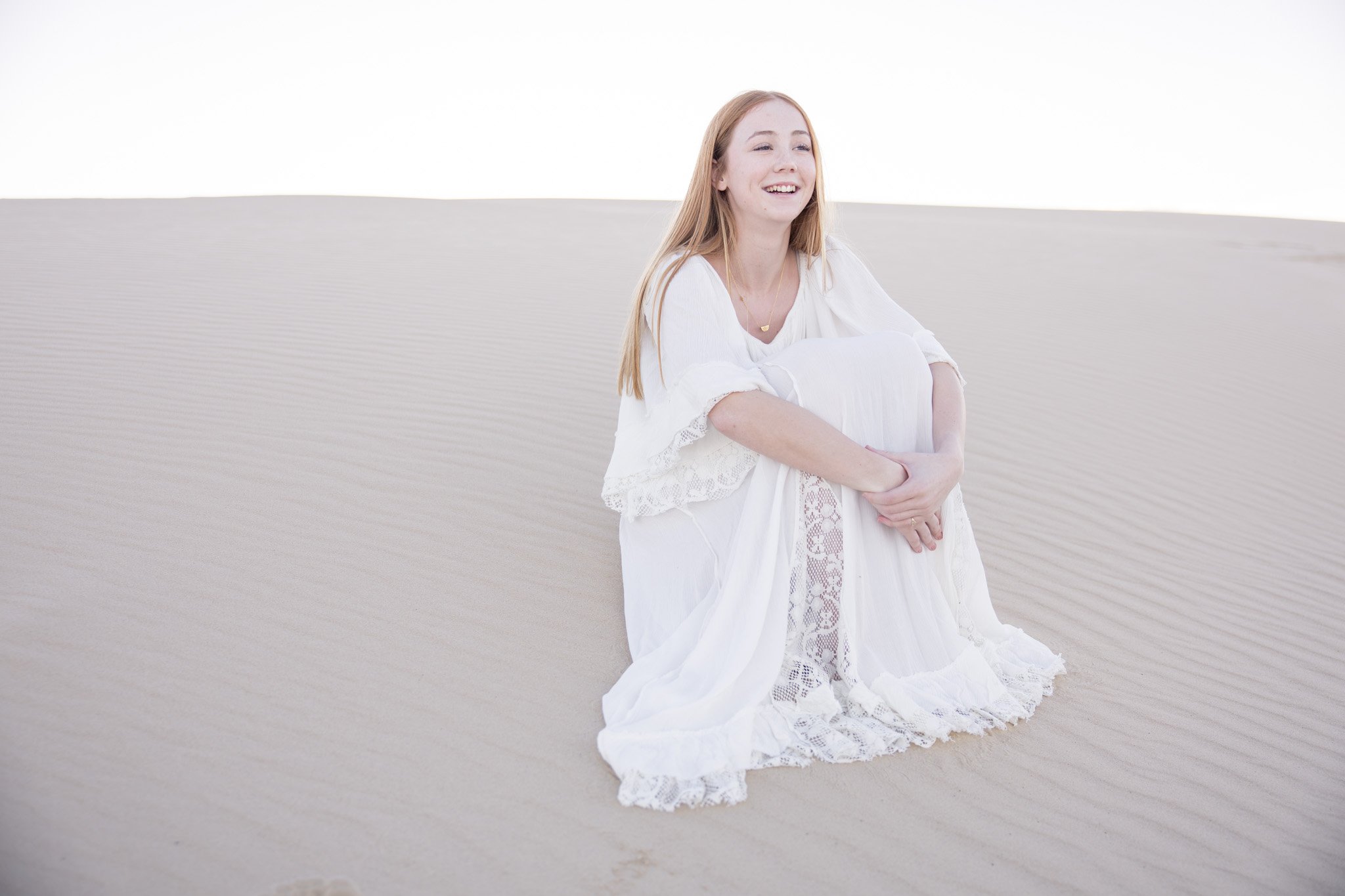 A young woman with long red hair in a white dress sitting on sand dunes, smiling with her arms crossed.