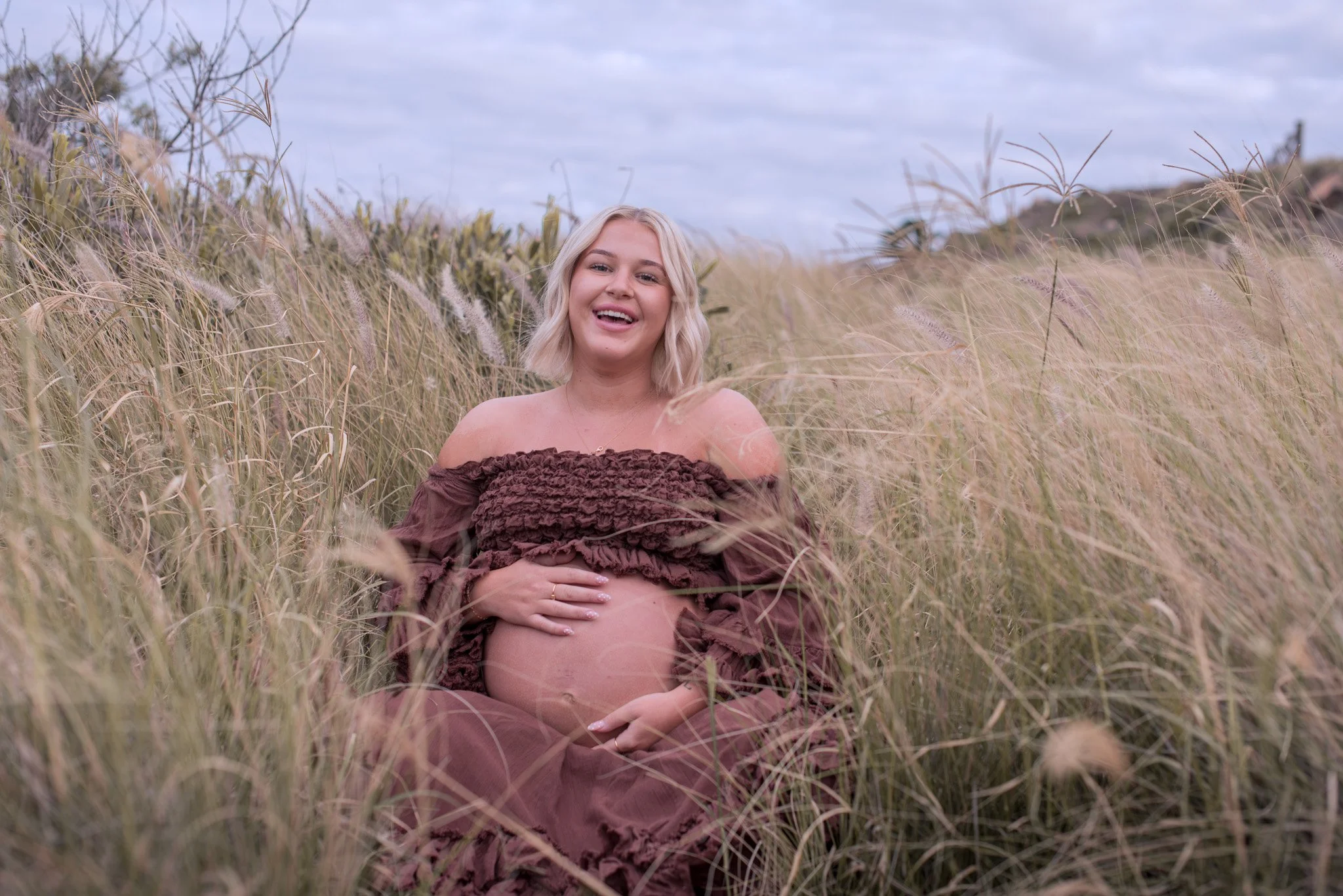 A pregnant woman with blonde hair sitting in a field of tall, dry grass, smiling at the camera.