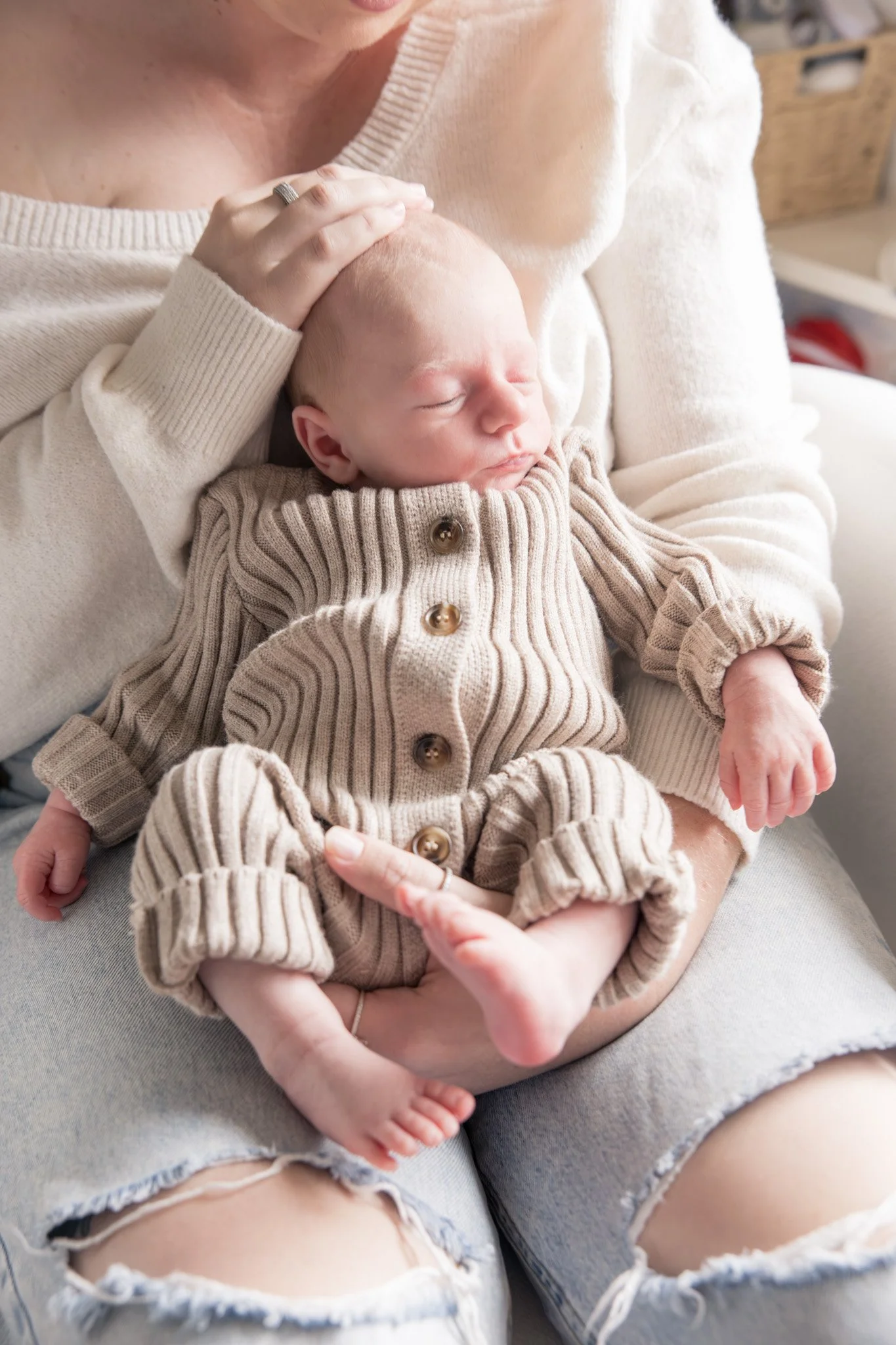 A woman holding a sleeping baby in her lap, with her hand resting on the baby's head. The woman is wearing a beige sweater, and the baby is dressed in a beige ribbed cardigan with wooden buttons. The woman is wearing ripped jeans.