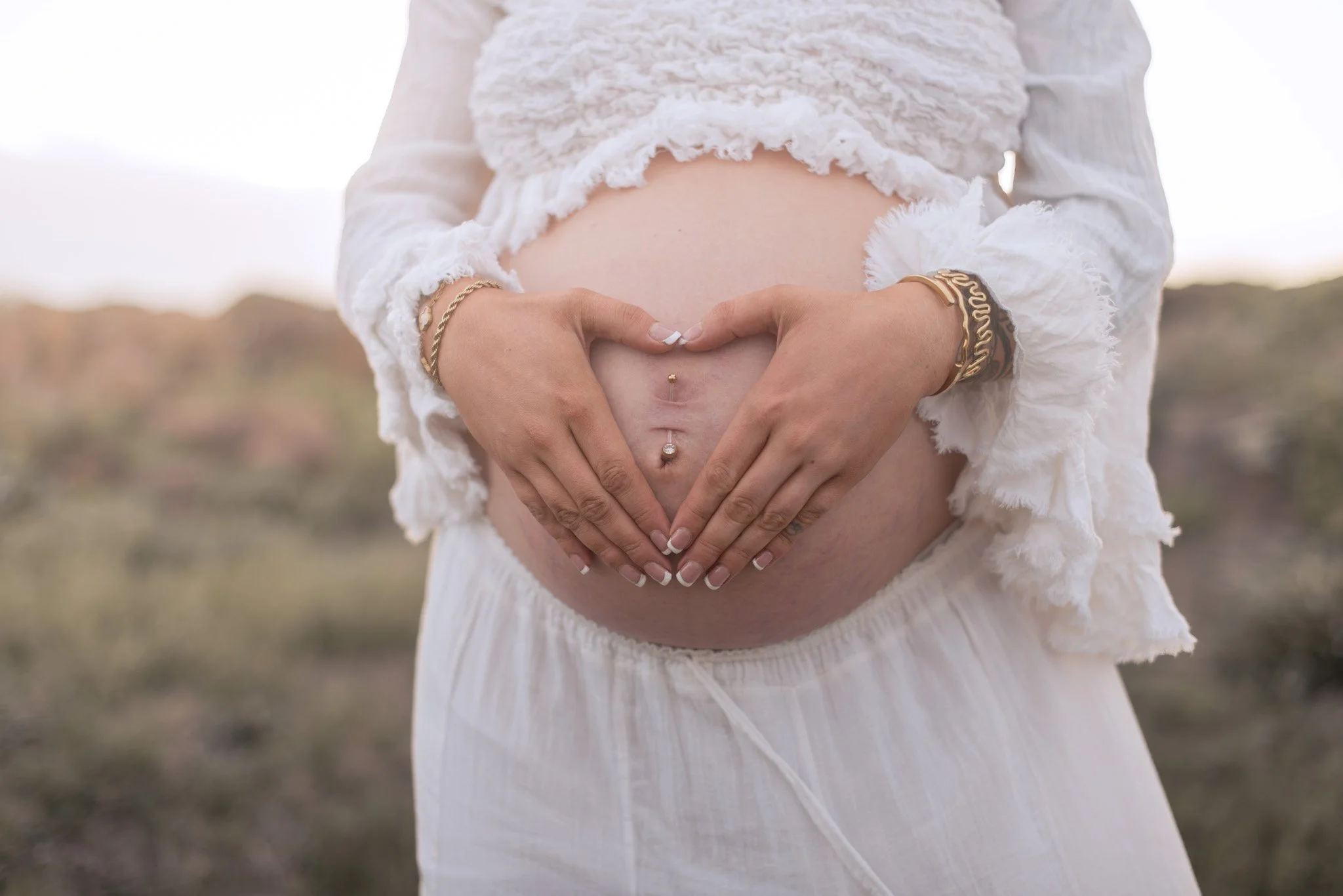 A pregnant woman holding her belly in a heart shape with her hands, wearing a white lace dress and jewelry, outdoors during sunset or sunrise. Port Stephens maternity session at anna bay beach