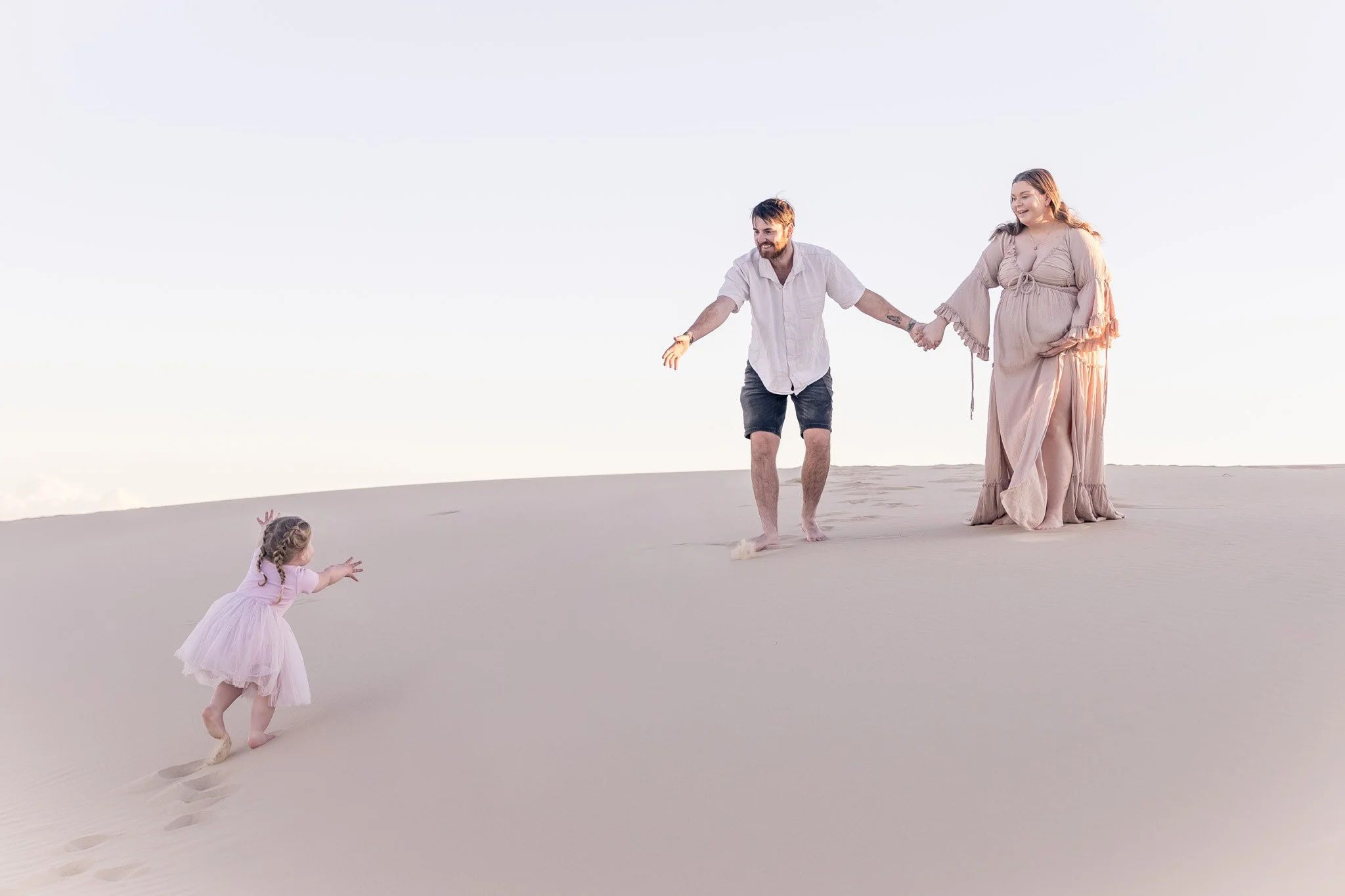 A couple holding hands walking on a sandy beach with two young girls playing in the sand. The sky is clear and the scene appears to be during sunset or sunrise.