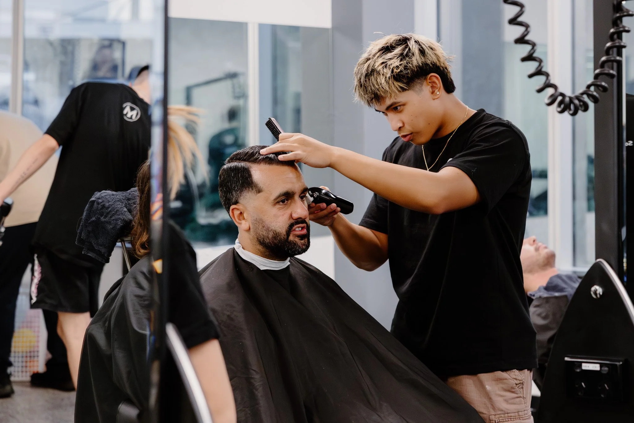 Barber trimming a man's hair at a barbershop while another customer is lying in a chair in the background.