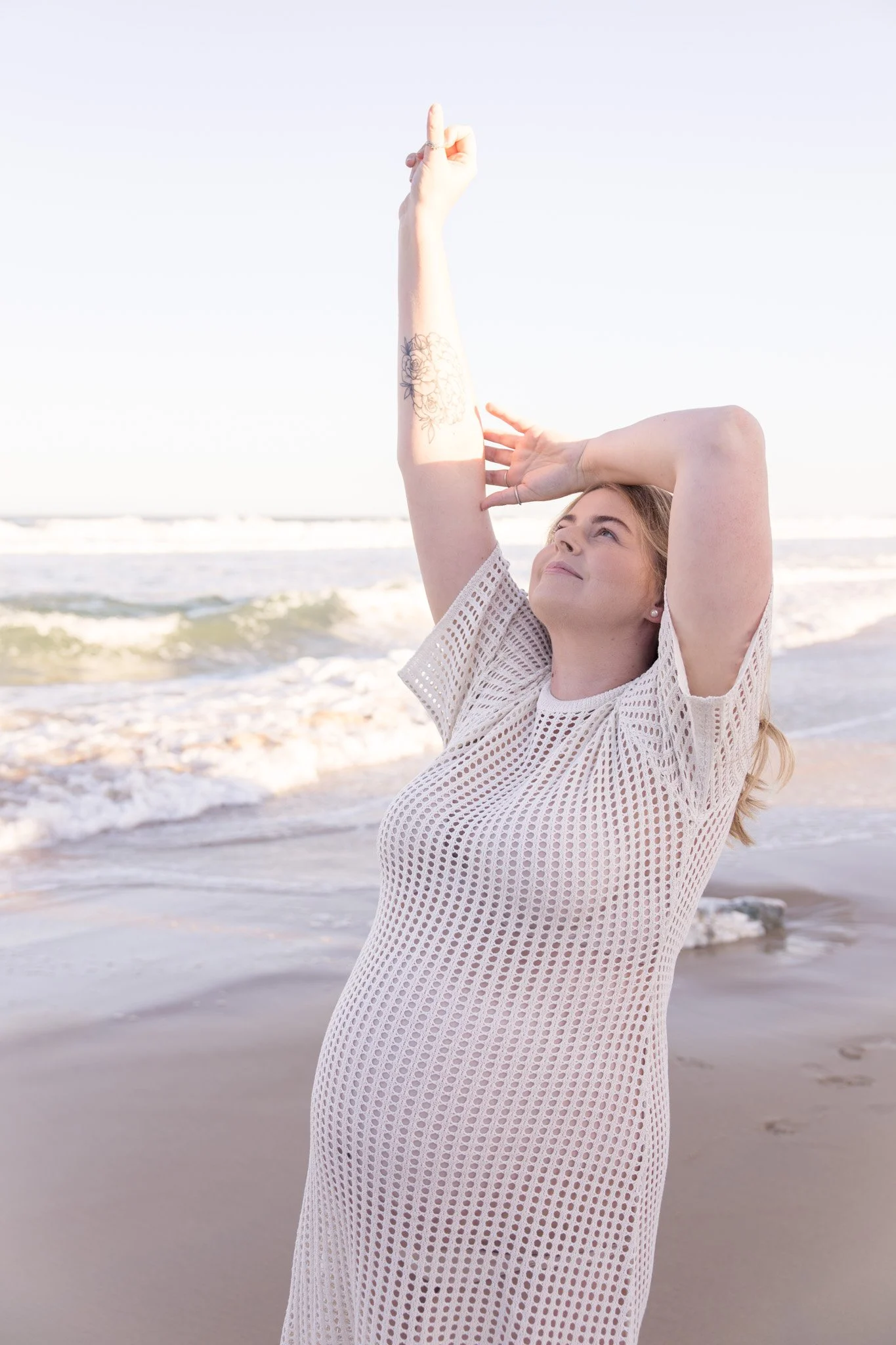 A woman with blonde hair and a floral tattoo on her forearm poses on a beach, looking upward with one arm raised and the other resting on her head, wearing a white mesh cover-up with ocean waves in the background.