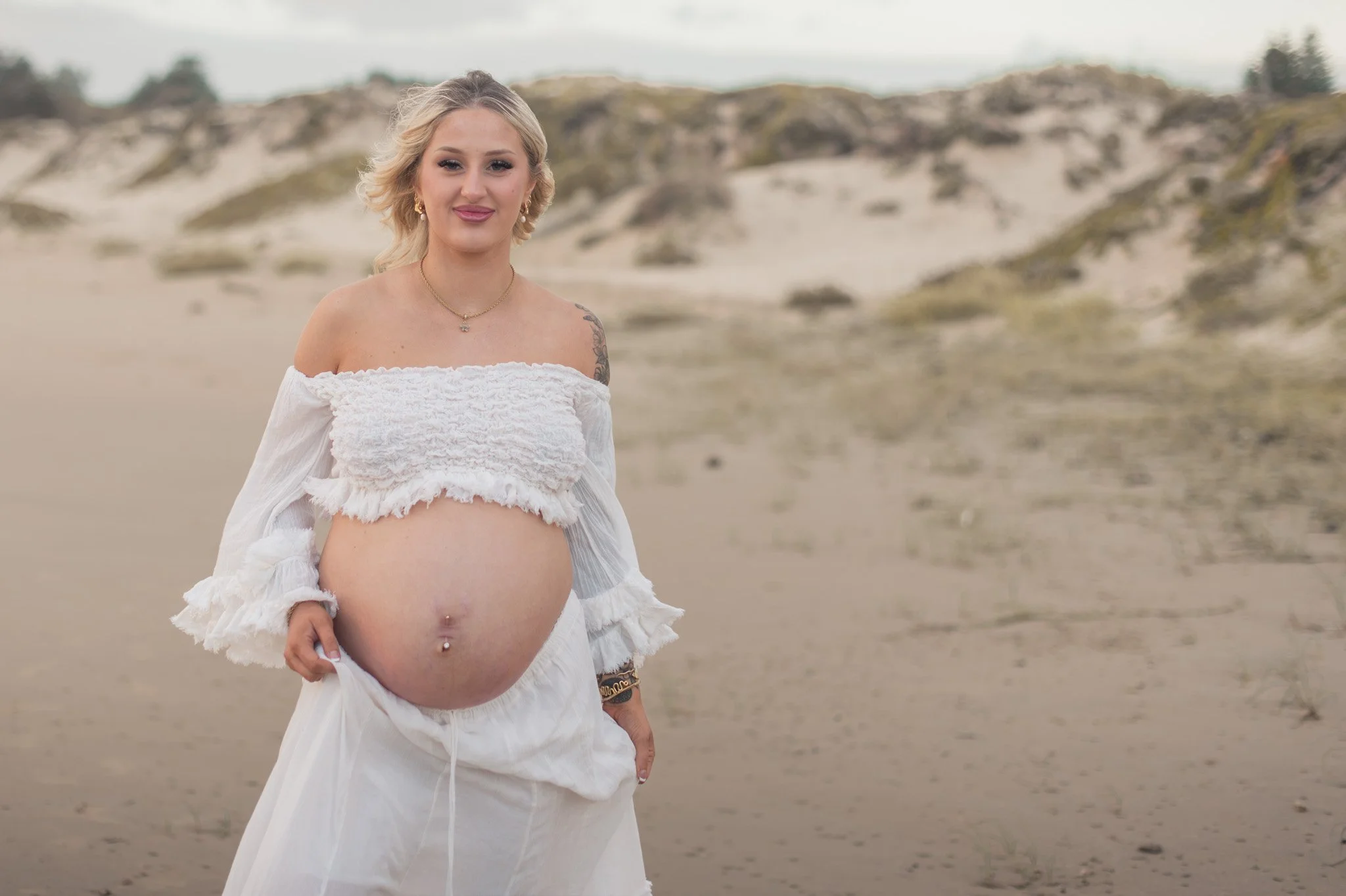 Pregnant woman standing on a beach with sand dunes in the background, wearing a white off-shoulder top and white skirt, smiling at the camera. During a pregnancy photoshoot at port stephens