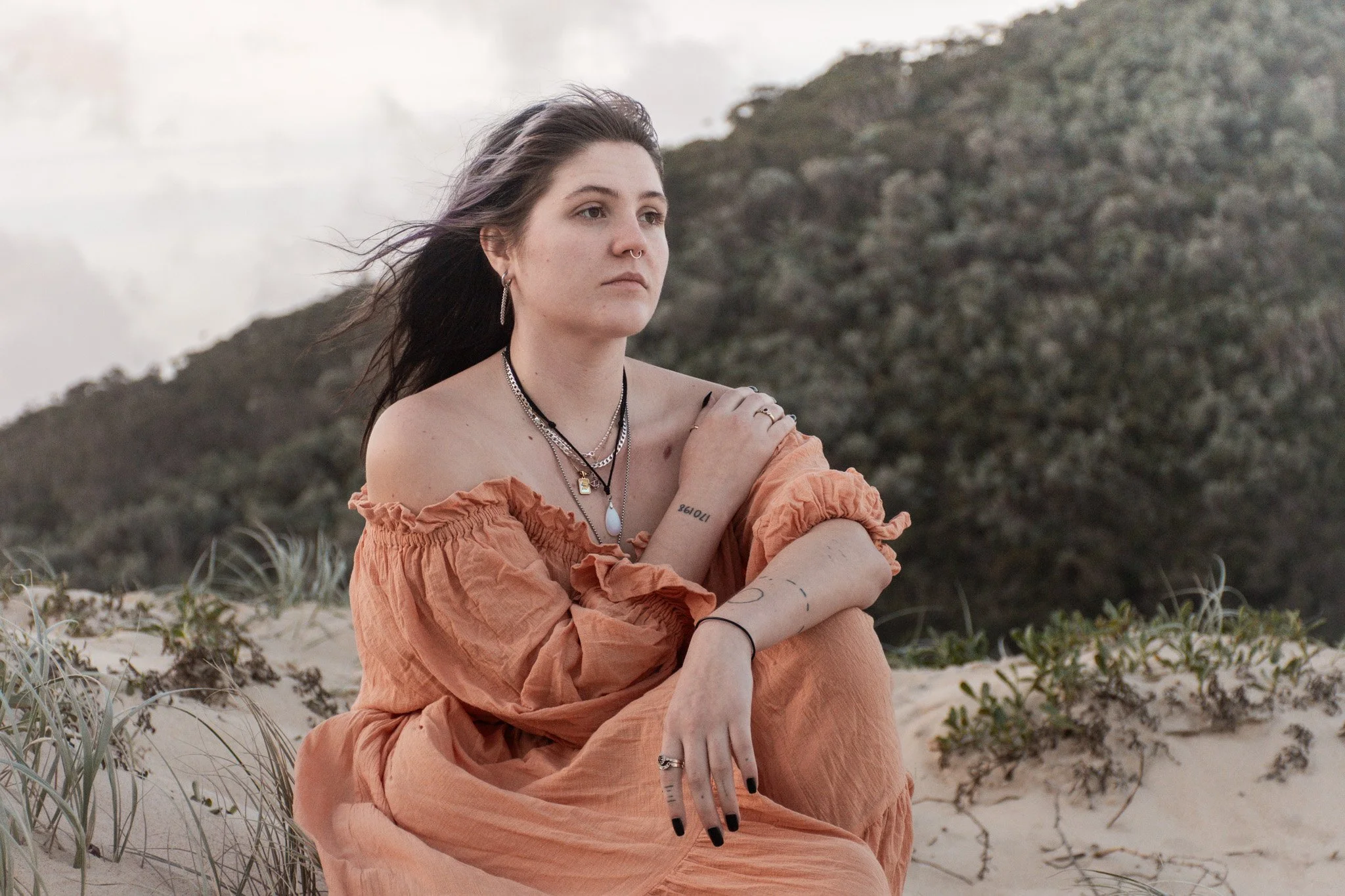Young woman with long brown hair, wearing layered necklaces and jewelry, sitting on a sandy and grassy area near mountains with a cloudy sky, dressed in an orange off-shoulder top.