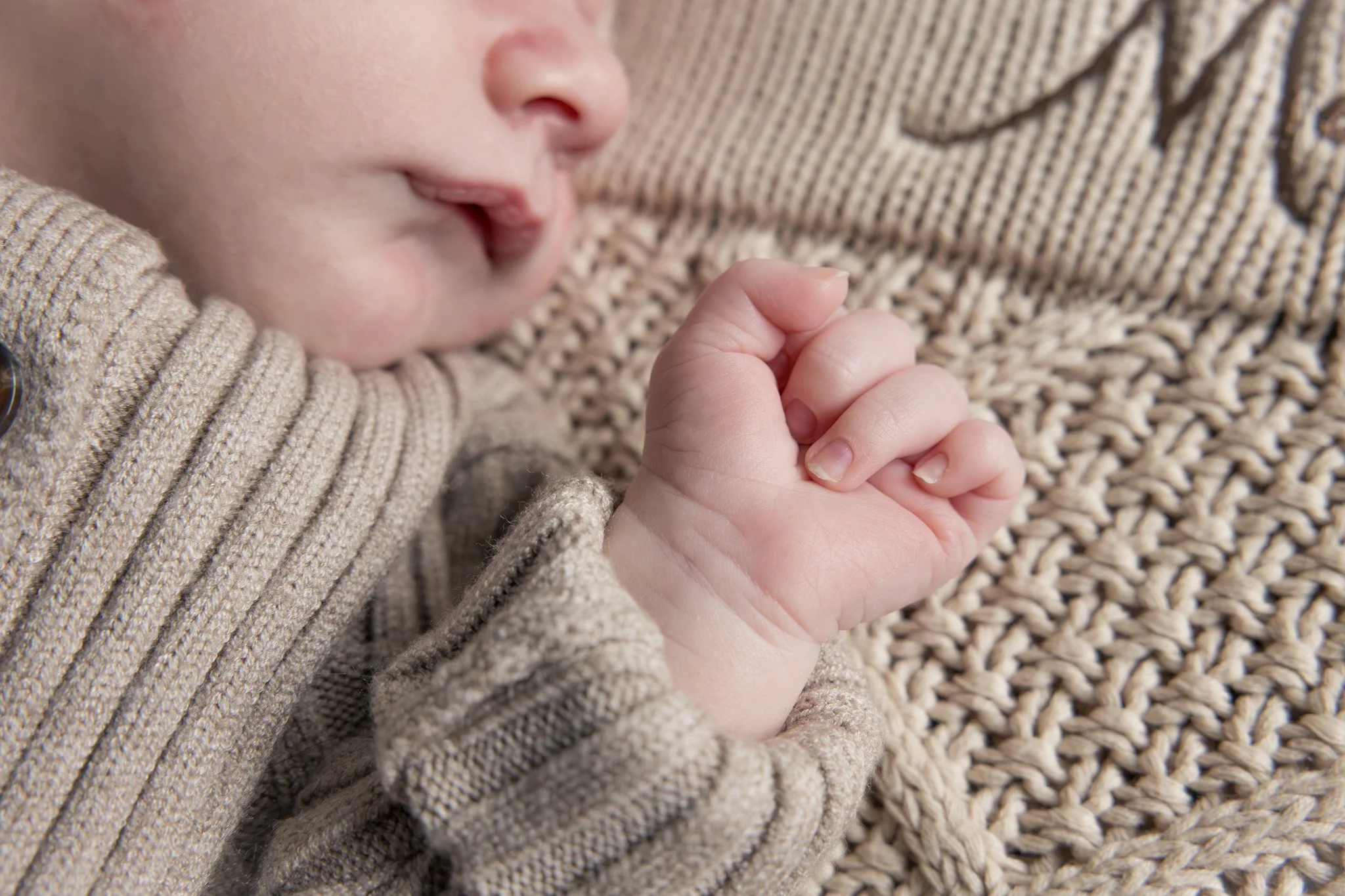 Close-up of a baby holding a person's finger, lying on a textured woven surface with a beige knitted blanket nearby.