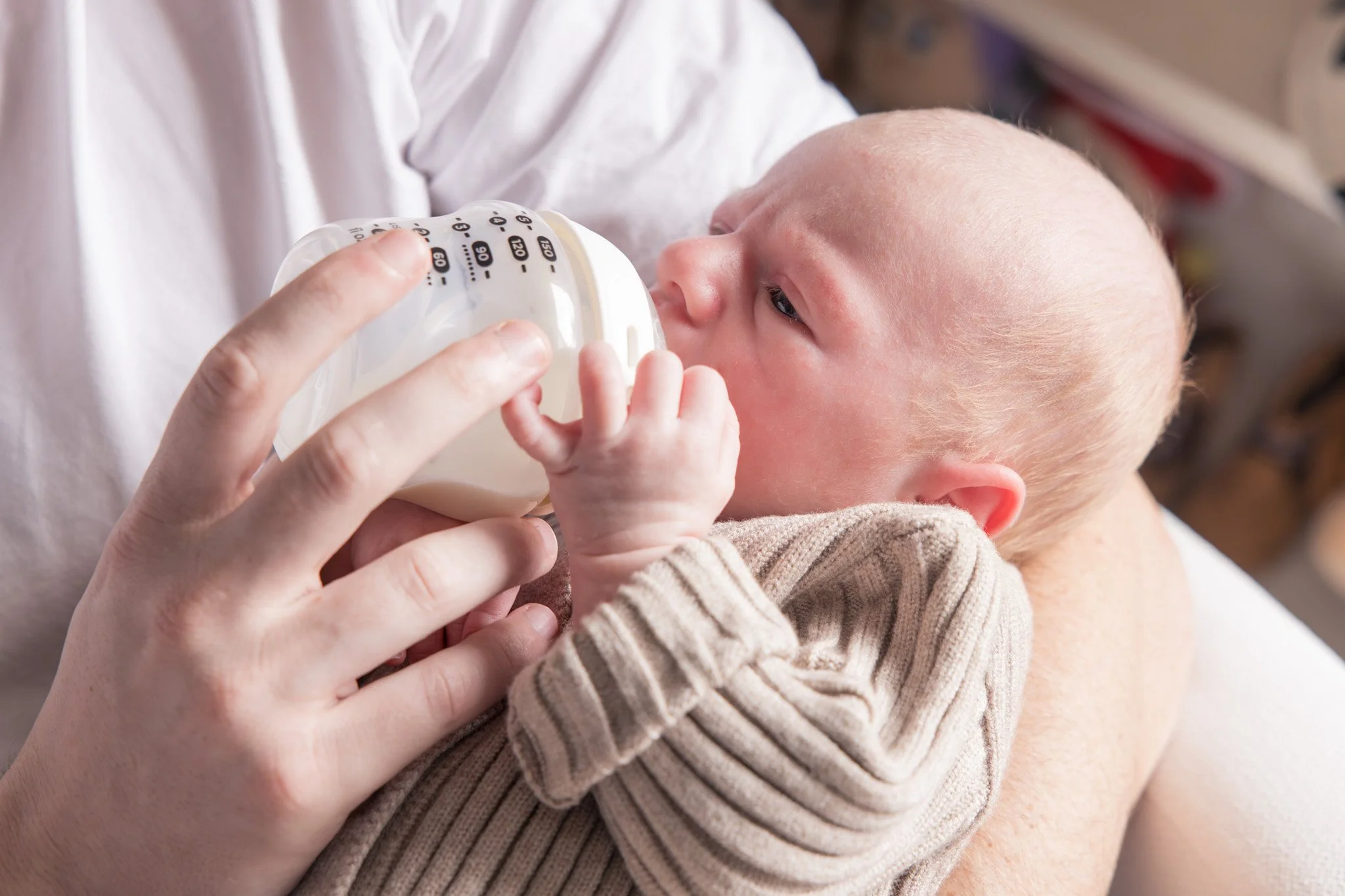Close-up of a baby being fed with a bottle by an adult, with the baby's face partially visible and the adult's hand holding the bottle.