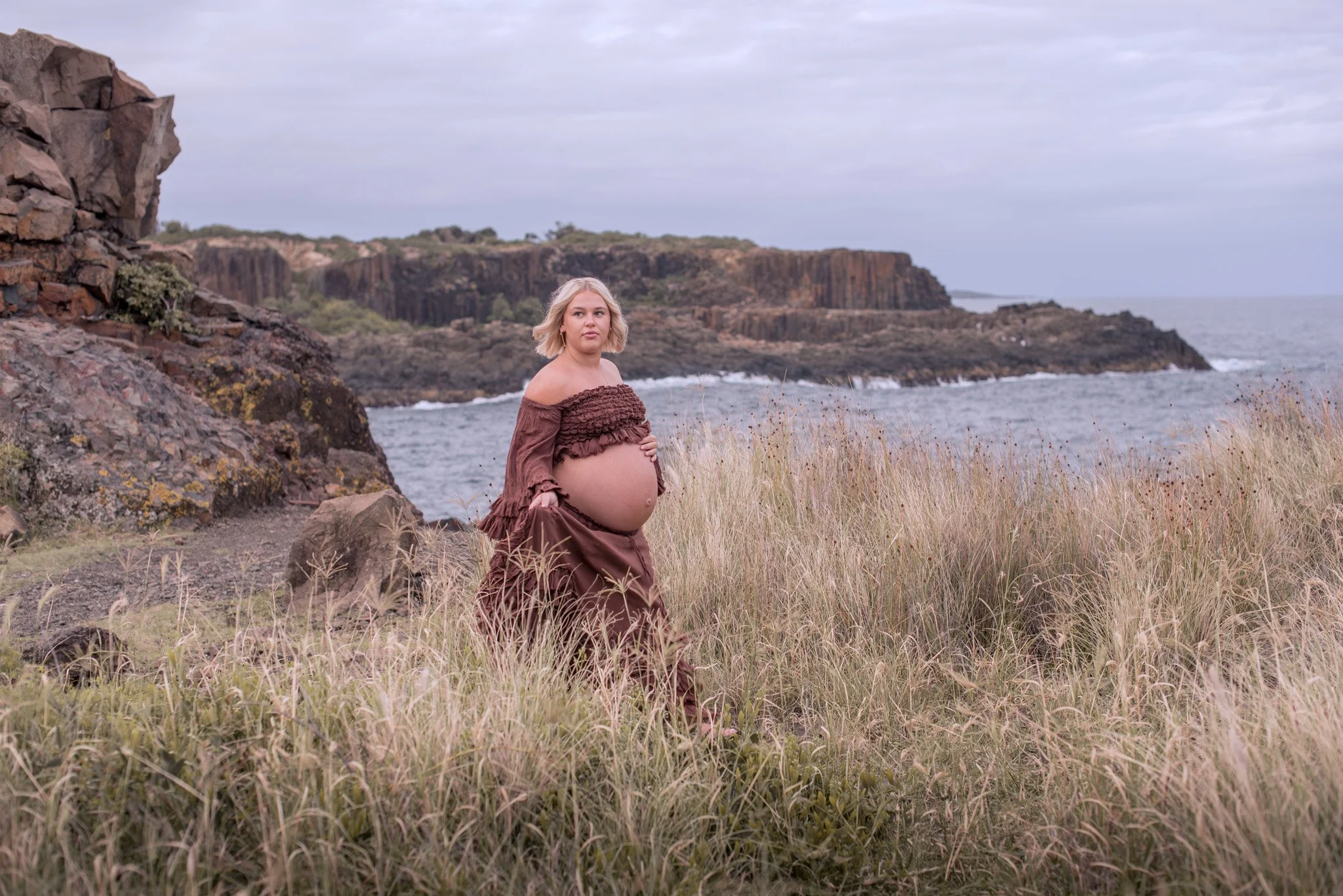 A pregnant woman stands on a grassy shoreline near rocky cliffs overlooking the ocean, wearing a brown off-shoulder top and skirt, with blonde hair and a calm expression.