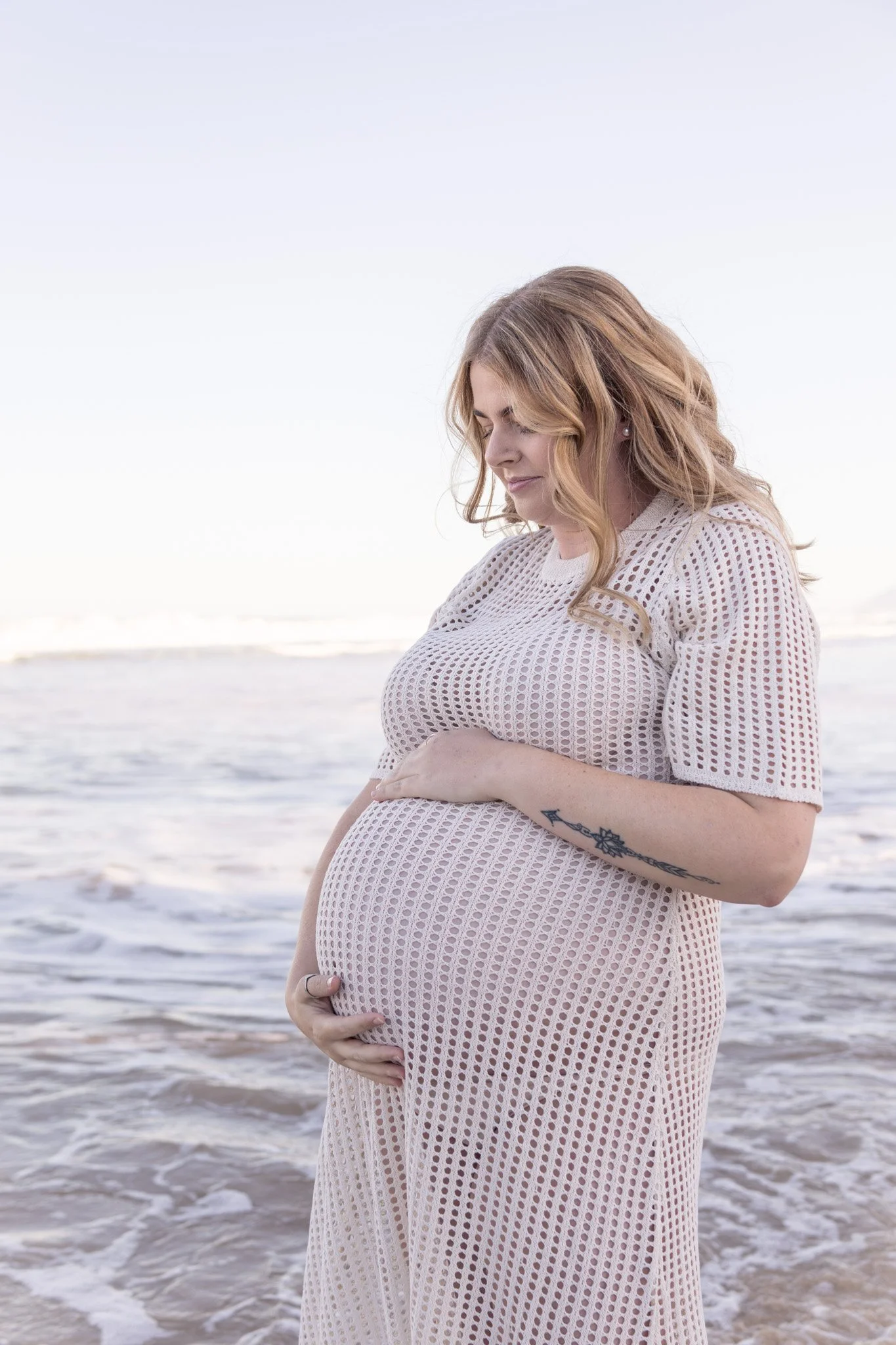 A pregnant woman with wavy blonde hair hugging her belly on the beach, wearing a white crochet dress.