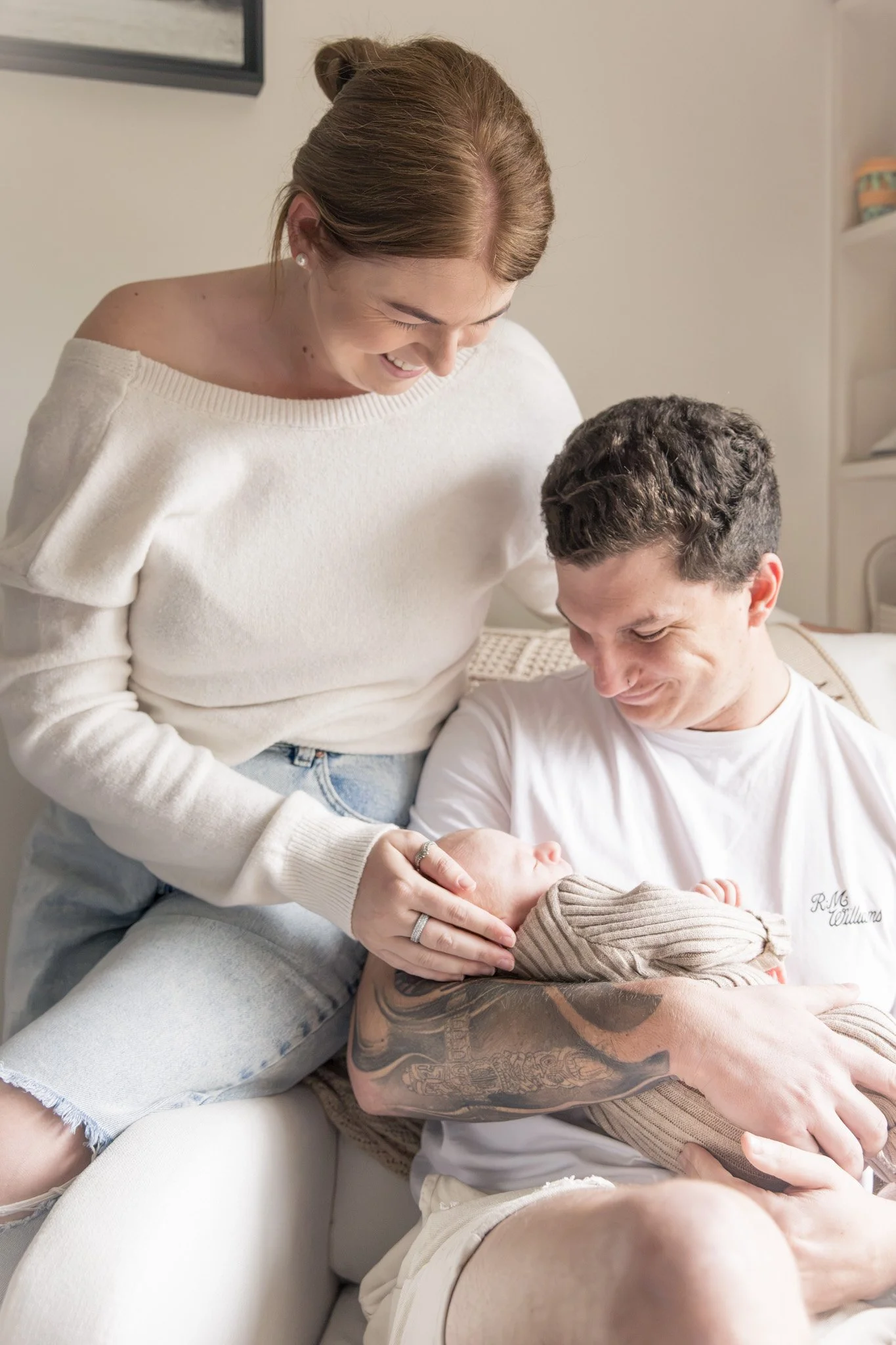 A woman smiling and looking down at a man with tattoos on his arm, who is holding a baby, all in a cozy indoor setting.