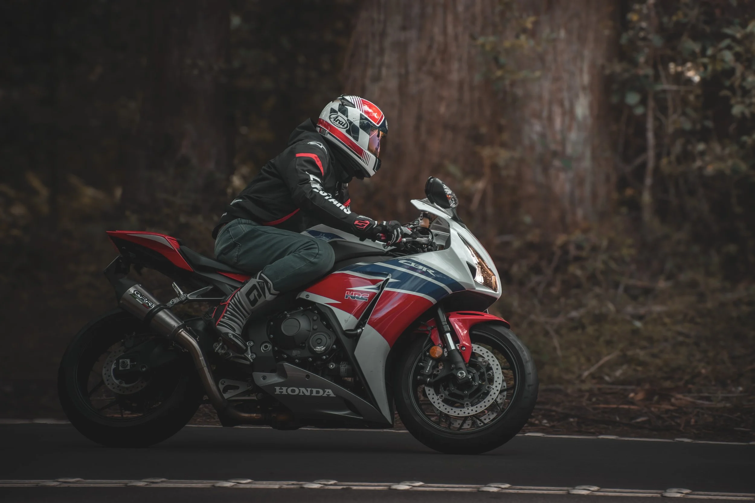 A person riding a red, white, and black Honda motorcycle along a paved road with trees in the background.