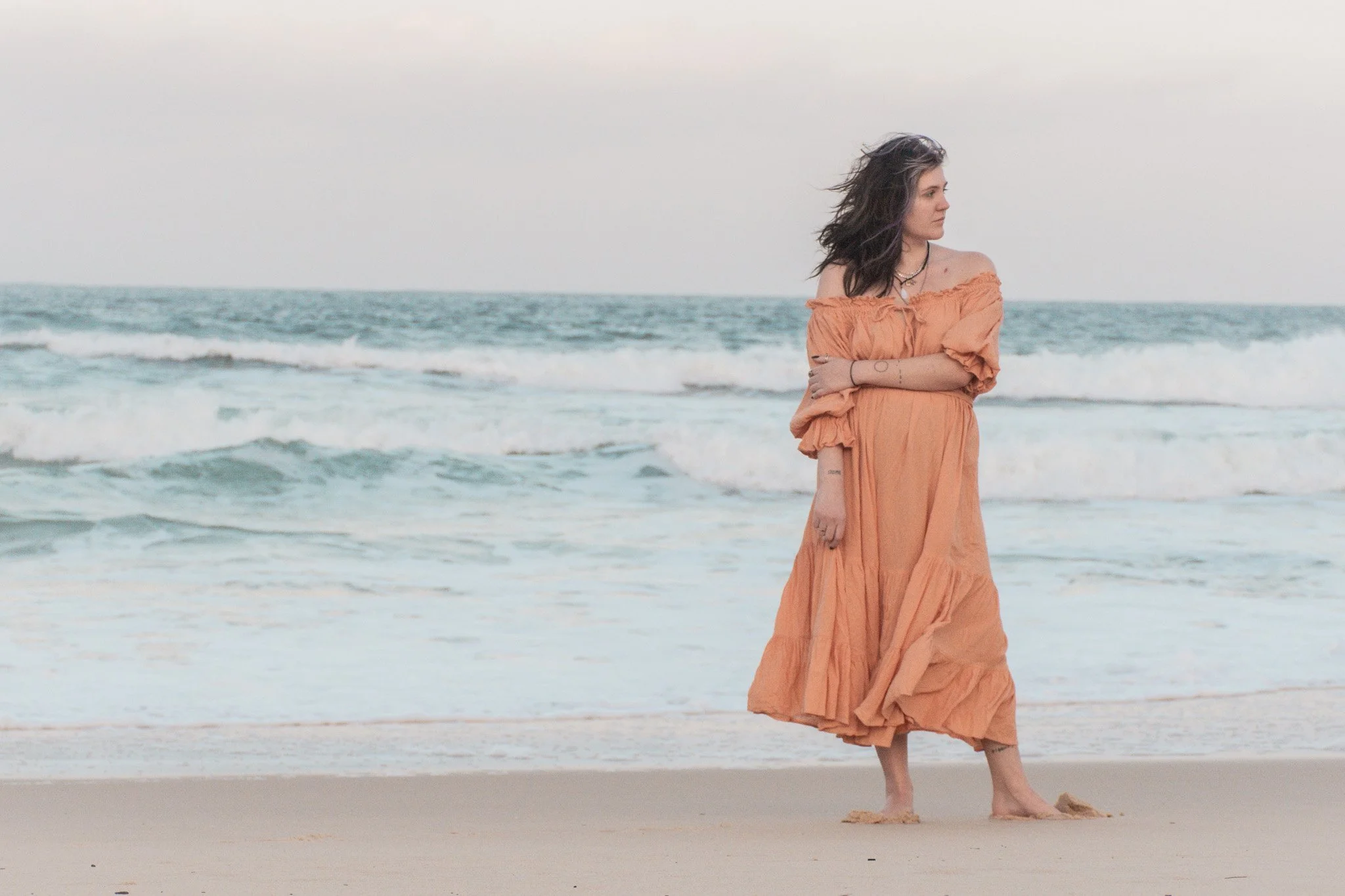 A woman with dark, wavy hair standing on the beach with ocean waves in the background. She is wearing an orange, off-the-shoulder dress and looking to the side with her arms crossed.