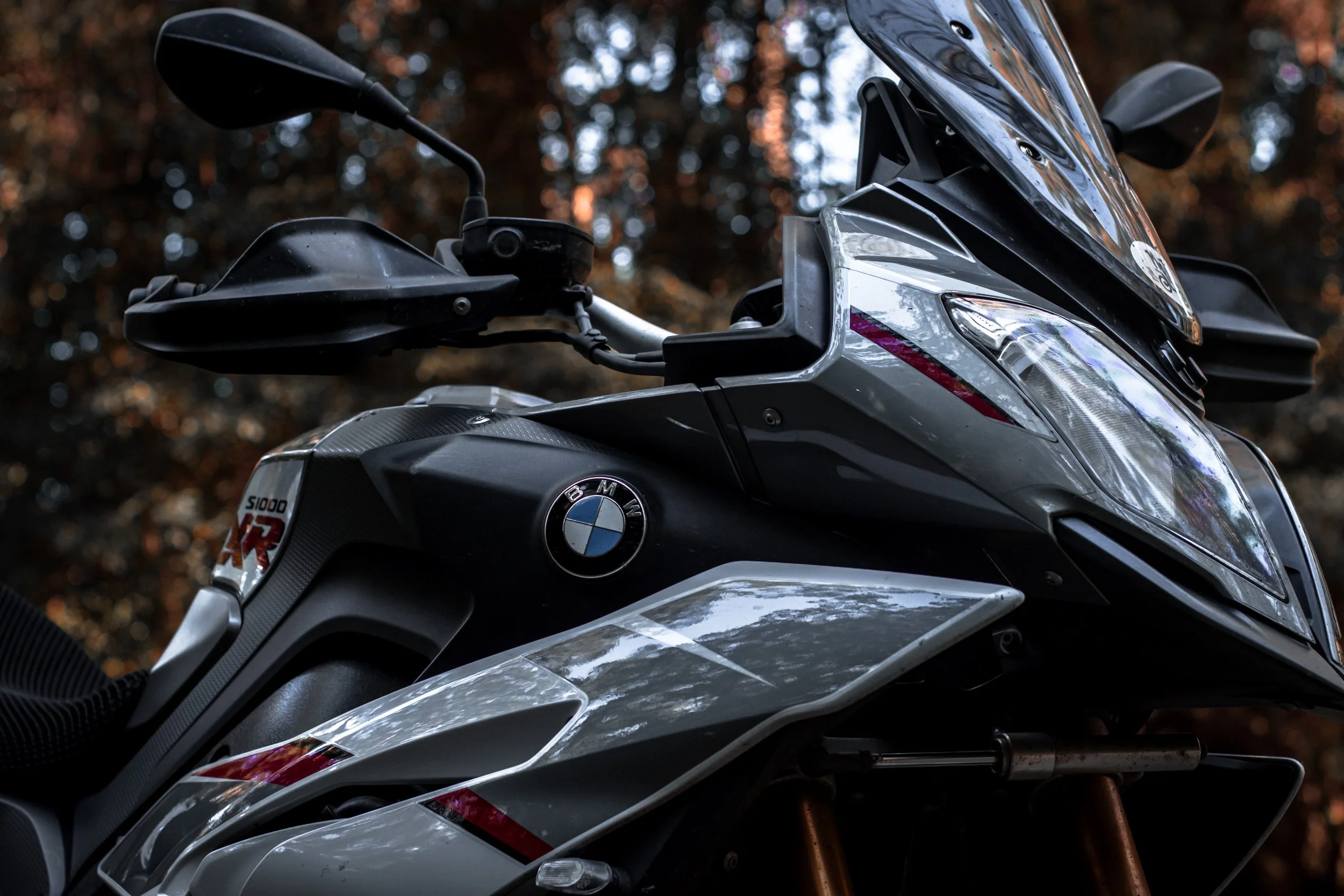Close-up of a BMW motorcycle with a BMW logo, fogged windshield, and black side mirrors against a blurry background of trees.