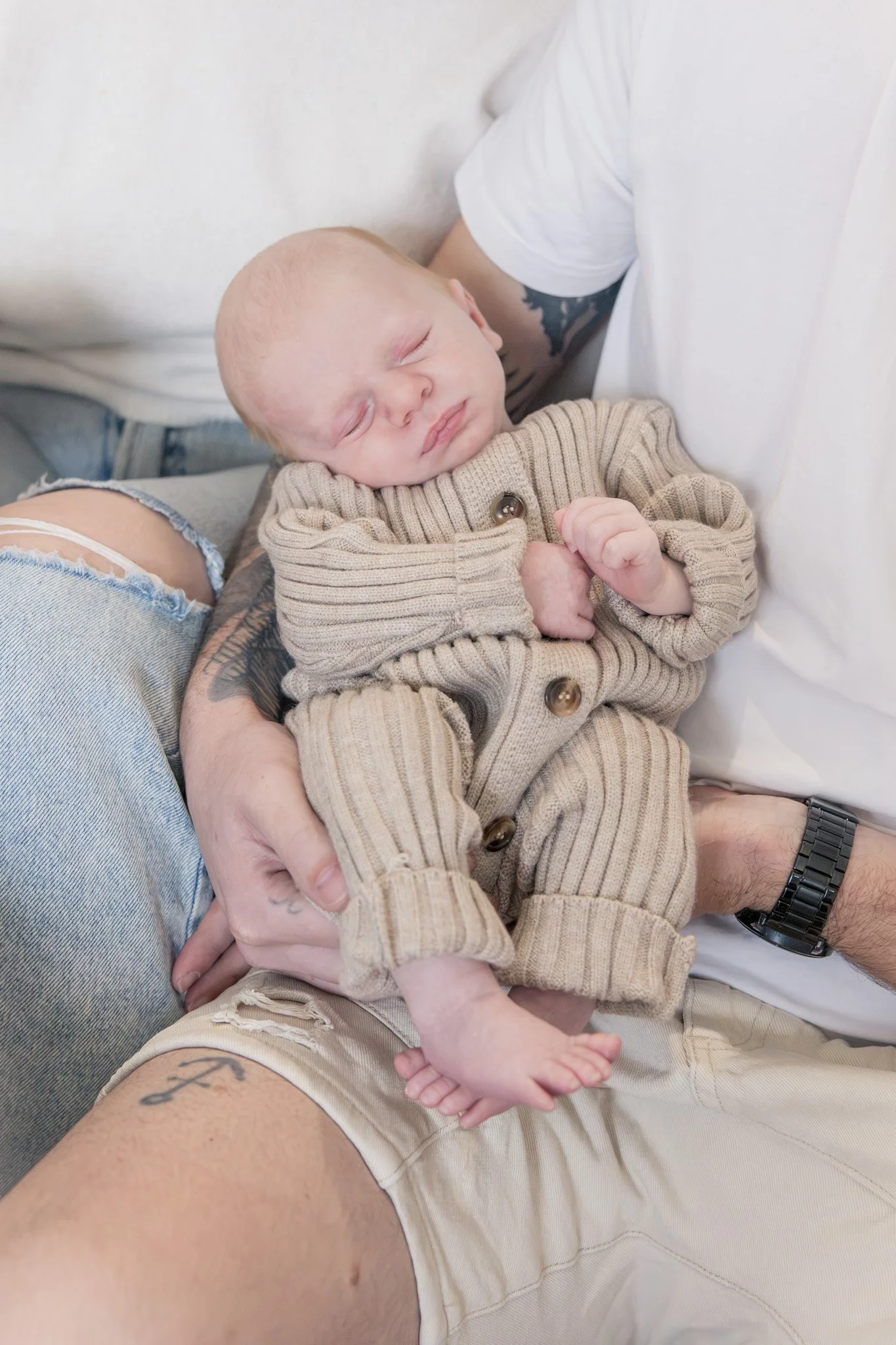 A person holding a sleeping baby on their lap, with the baby resting on the person's arm and the person wearing light-colored shorts and a white T-shirt.